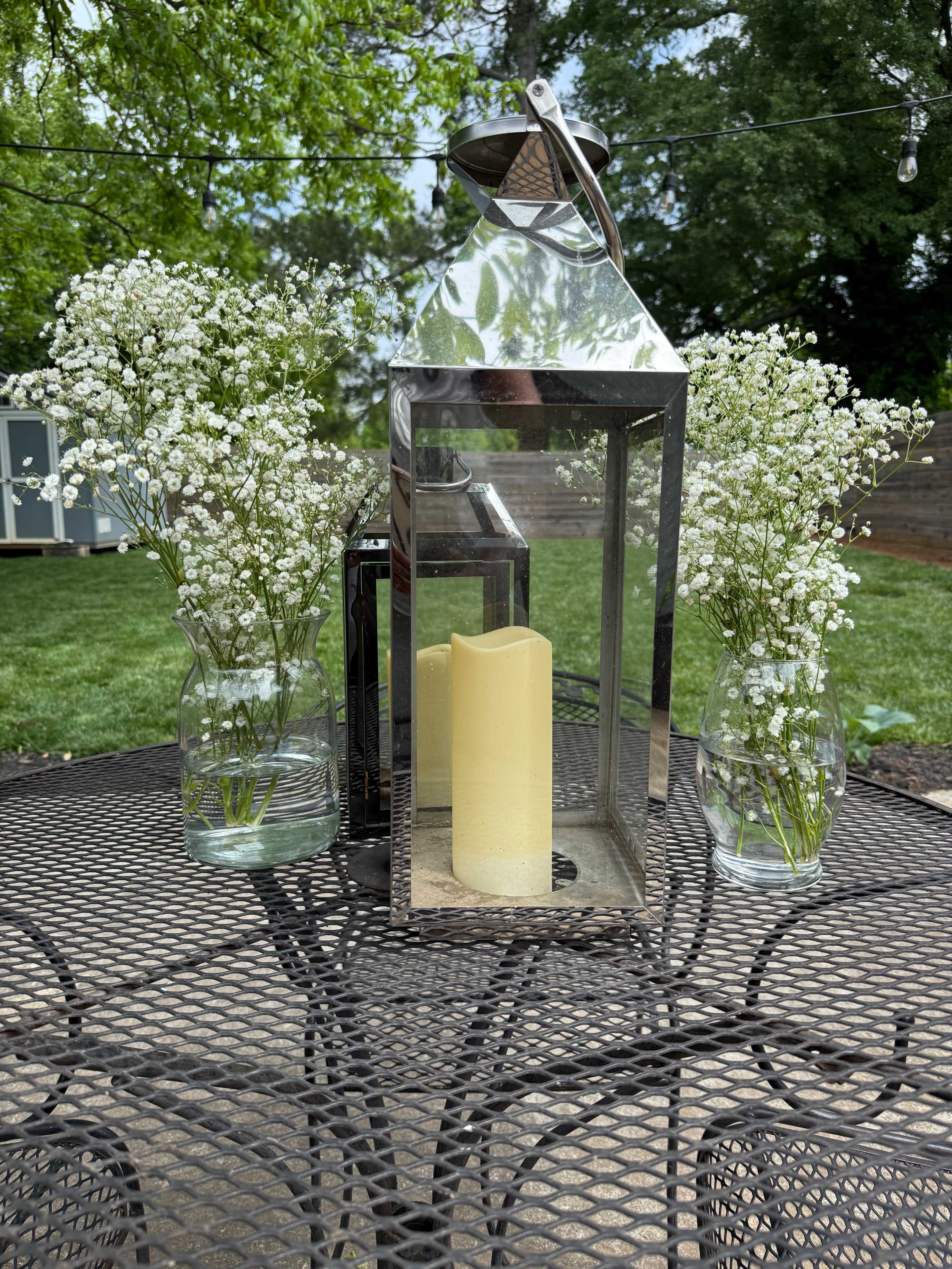 A glass lantern with a yellow candle is placed on a metal table surrounded by two vases of white flowers in a green backyard setting.