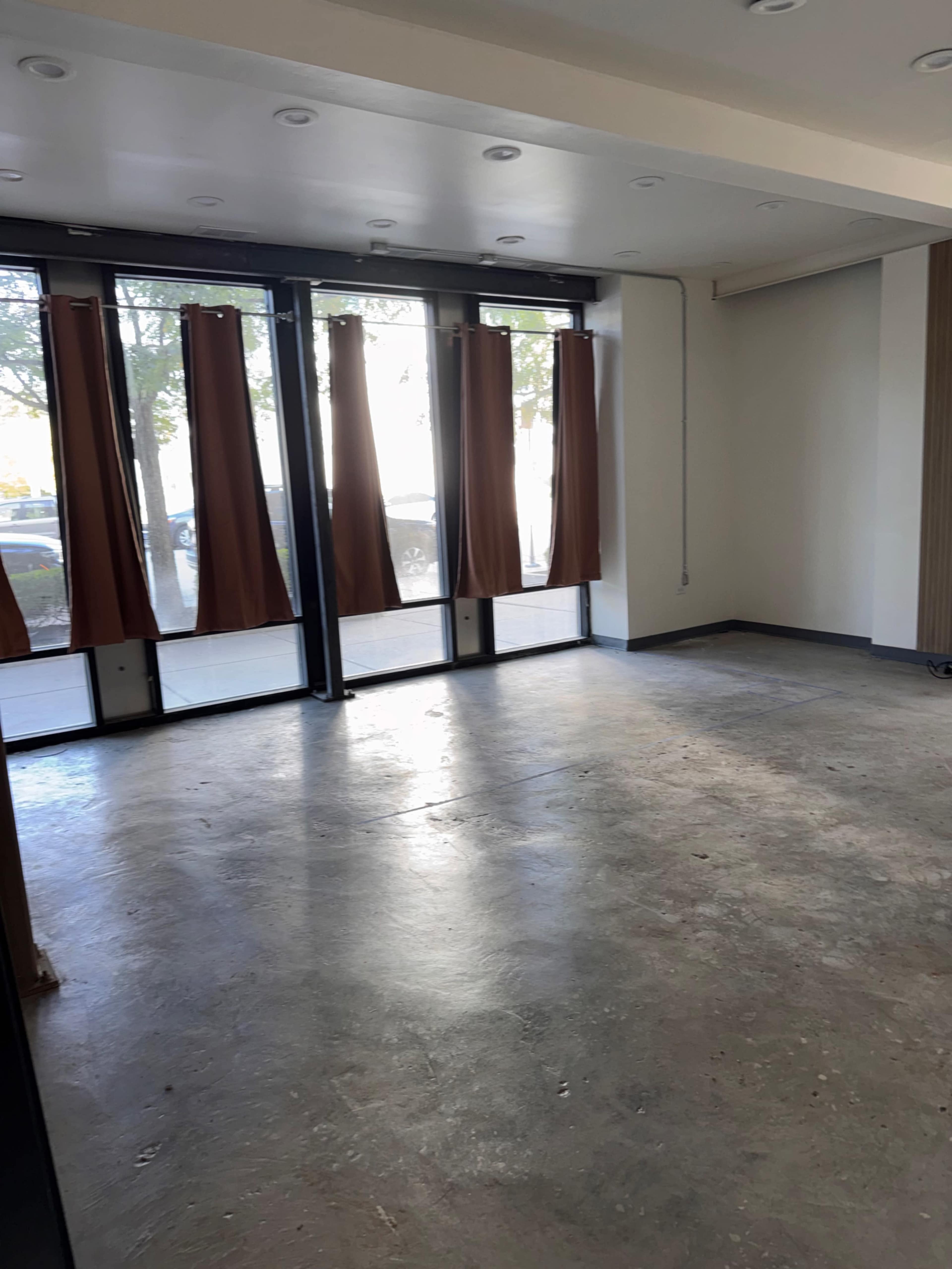 The image shows an empty room with concrete flooring, large windows covered by brown curtains, and a lack of furniture or decorations.