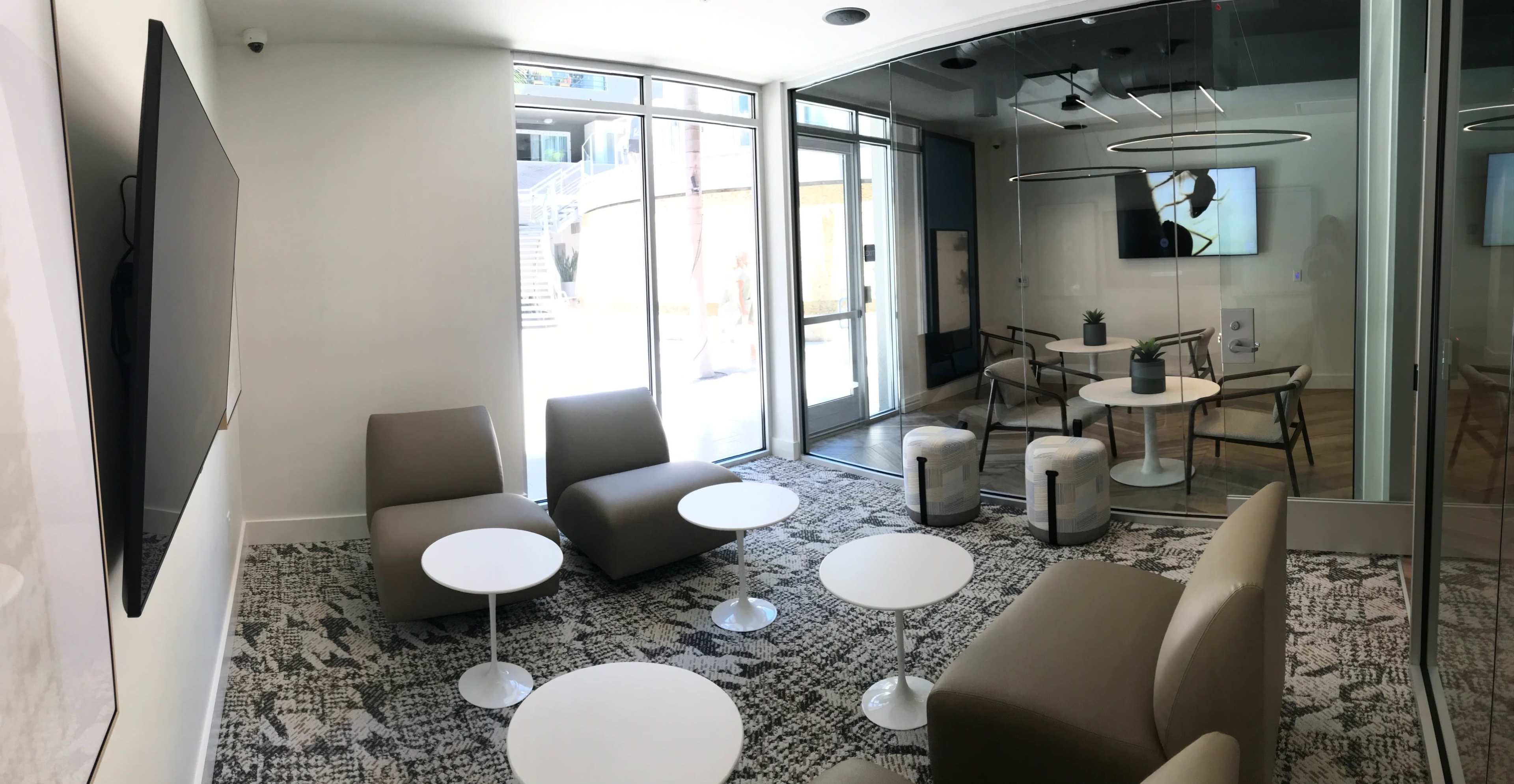 A modern lounge area with two seating arrangements, small round tables, and large glass doors leading to an outdoor space.