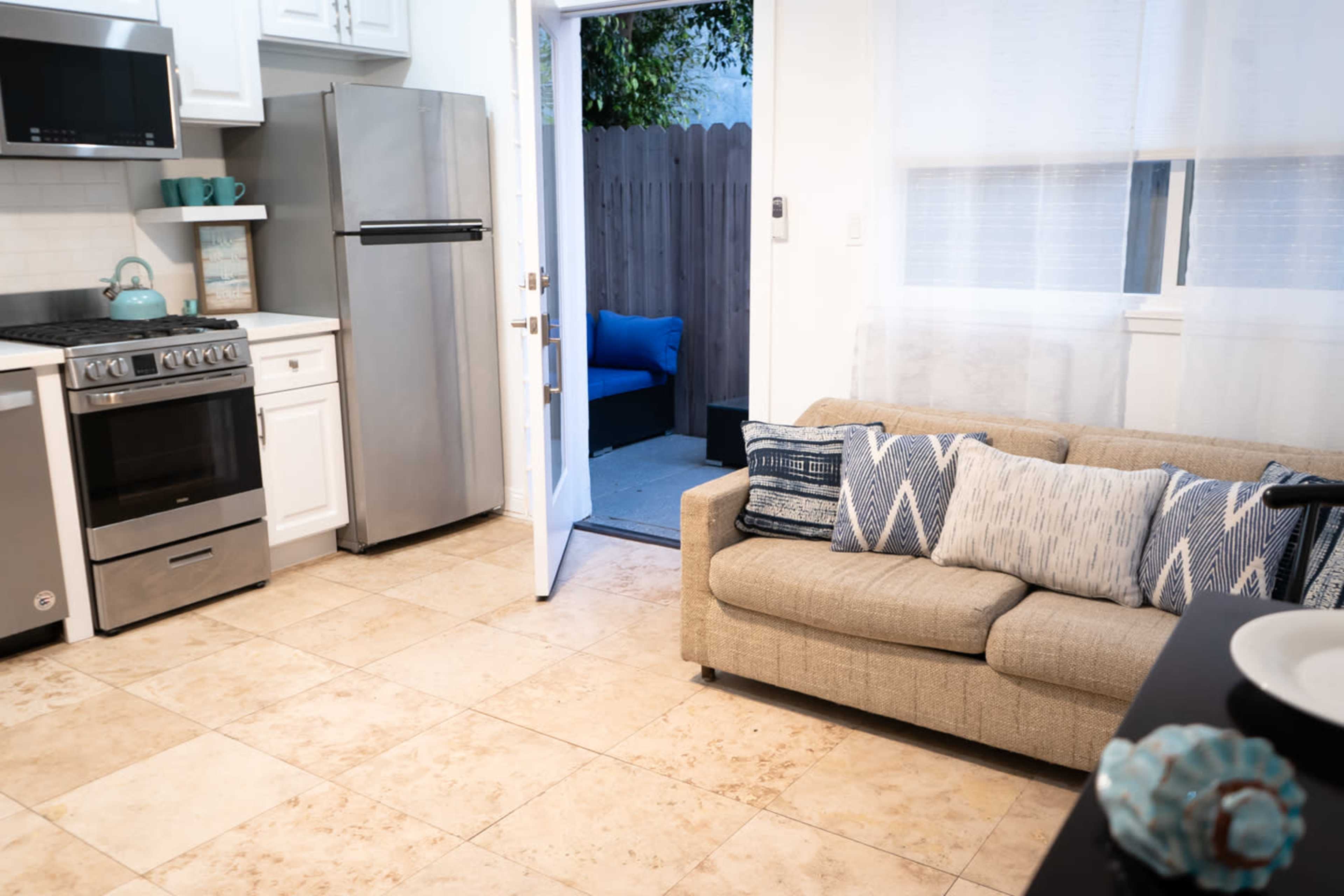 The image shows a kitchen area with a stainless steel refrigerator, oven, and a beige sofa with decorative pillows, leading to an outdoor seating area visible through an open door.
