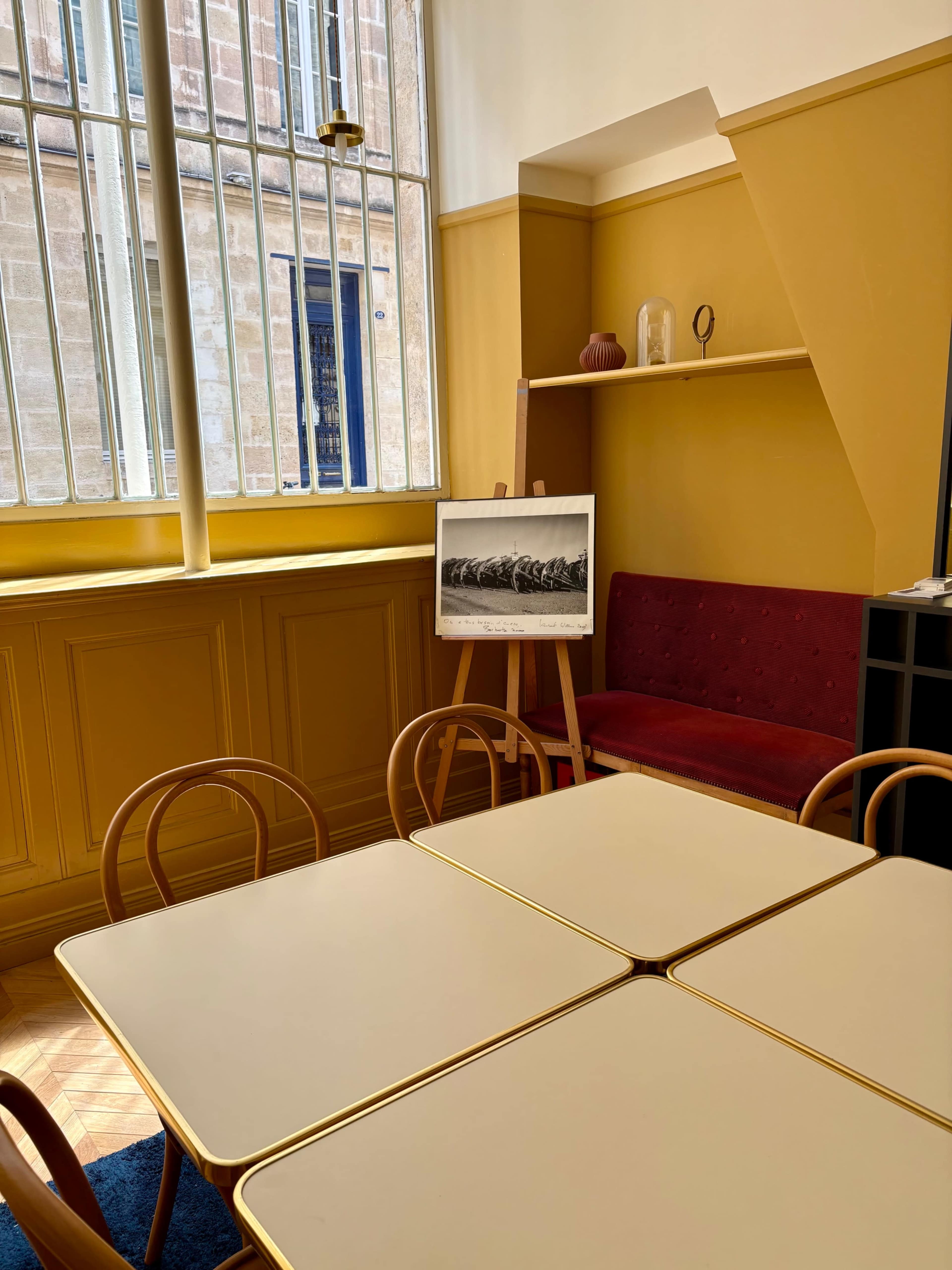 A bright room features a yellow wall, a red sofa, a wooden easel displaying a black-and-white photograph, and four tables with golden edges.