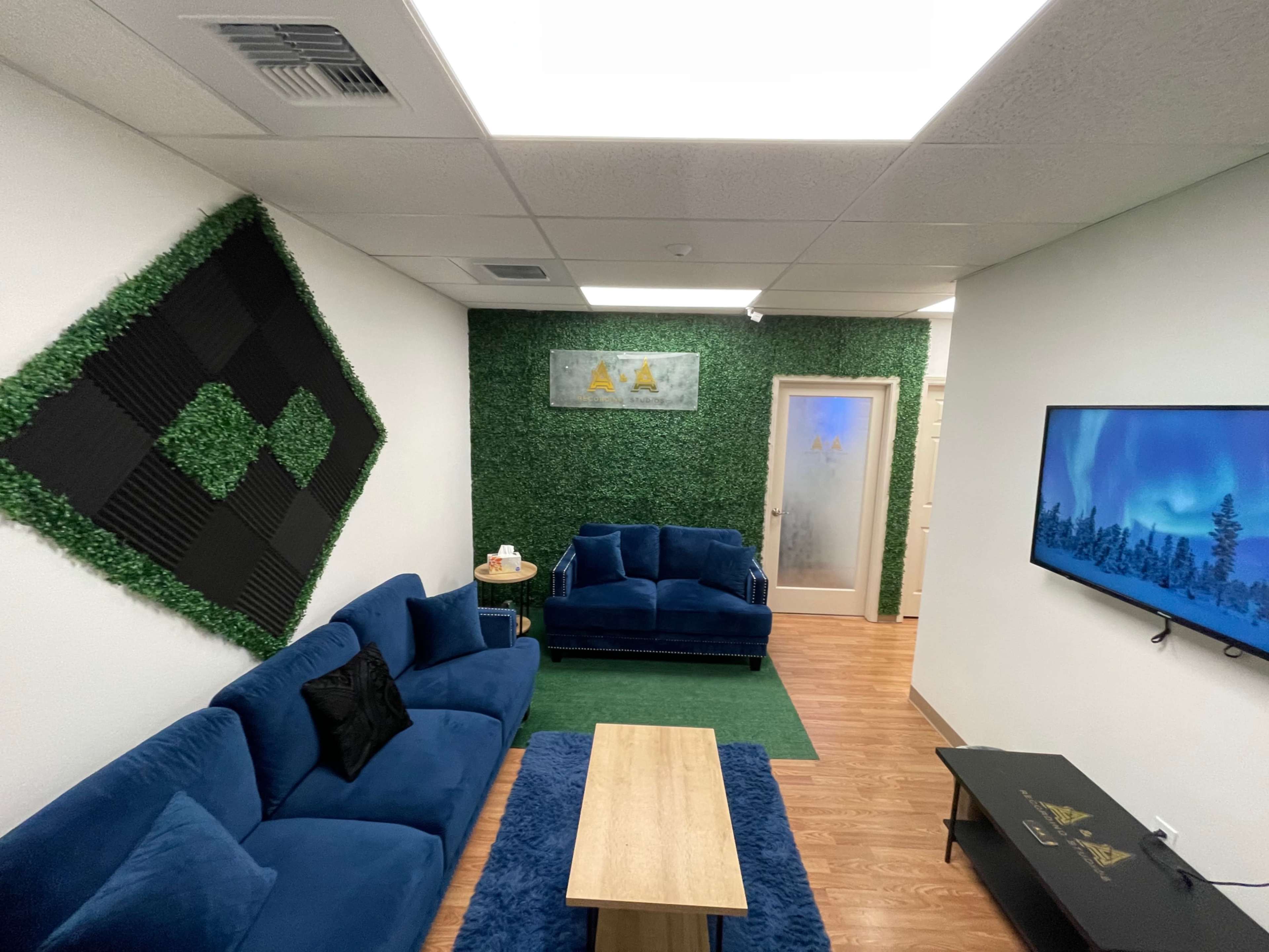 A modern lounge area with blue sofas, a wooden coffee table, artificial greenery on the walls, and a television mounted on one side.