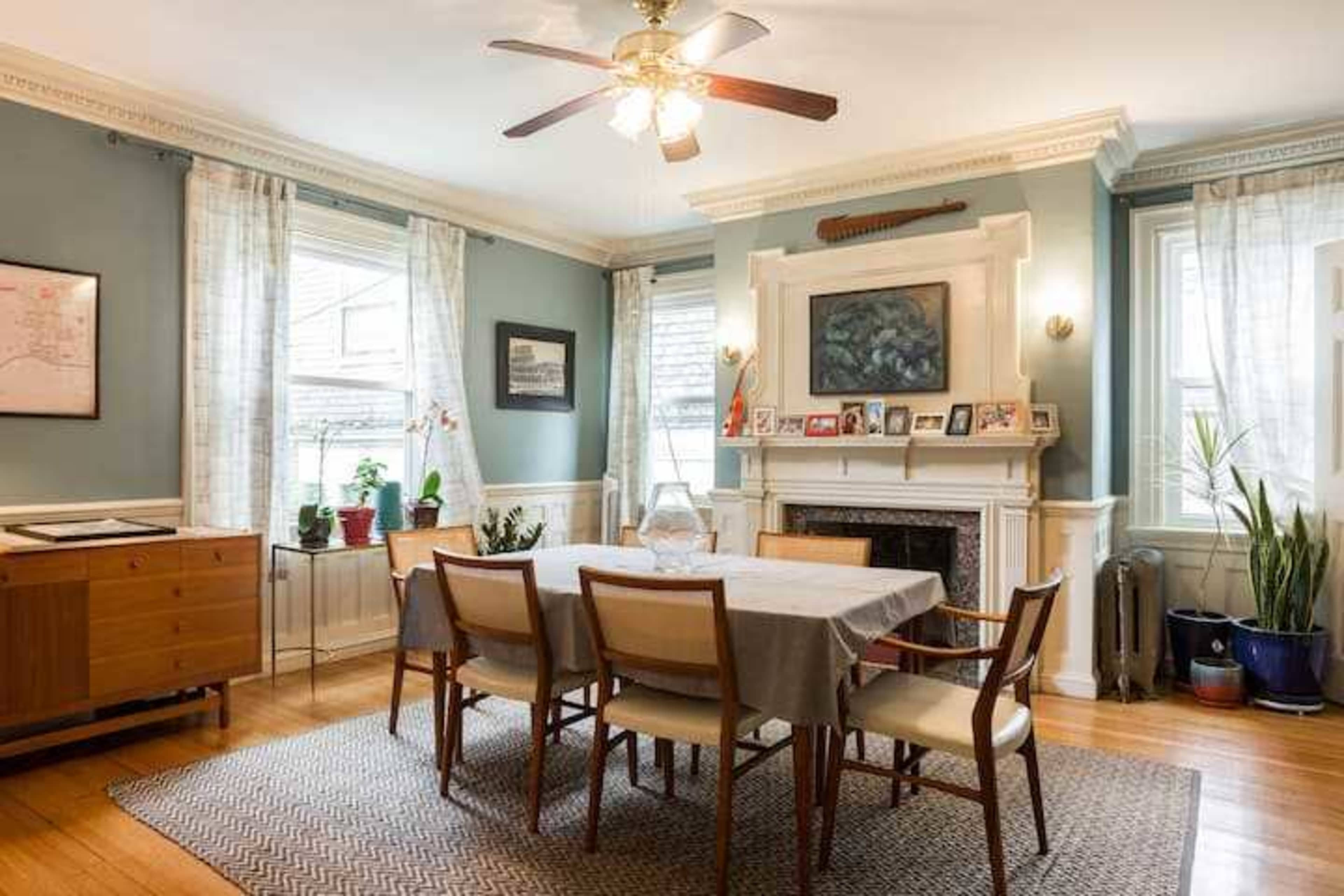A dining room features a rectangular table surrounded by chairs, with a fireplace and large windows adorned with light curtains.