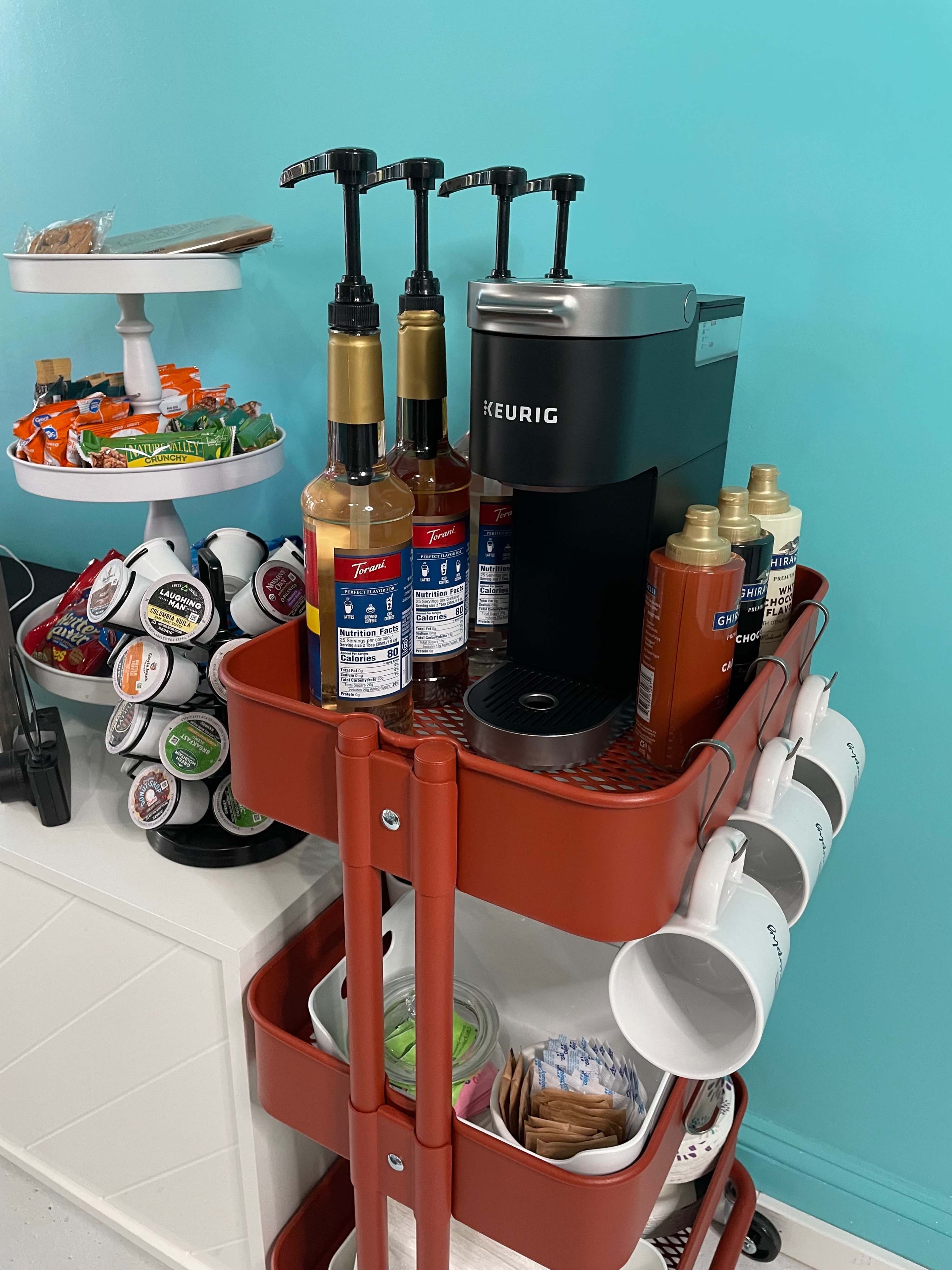 A coffee cart features a Keurig machine, flavored syrups, and a selection of coffee pods, along with mugs on the bottom shelf.