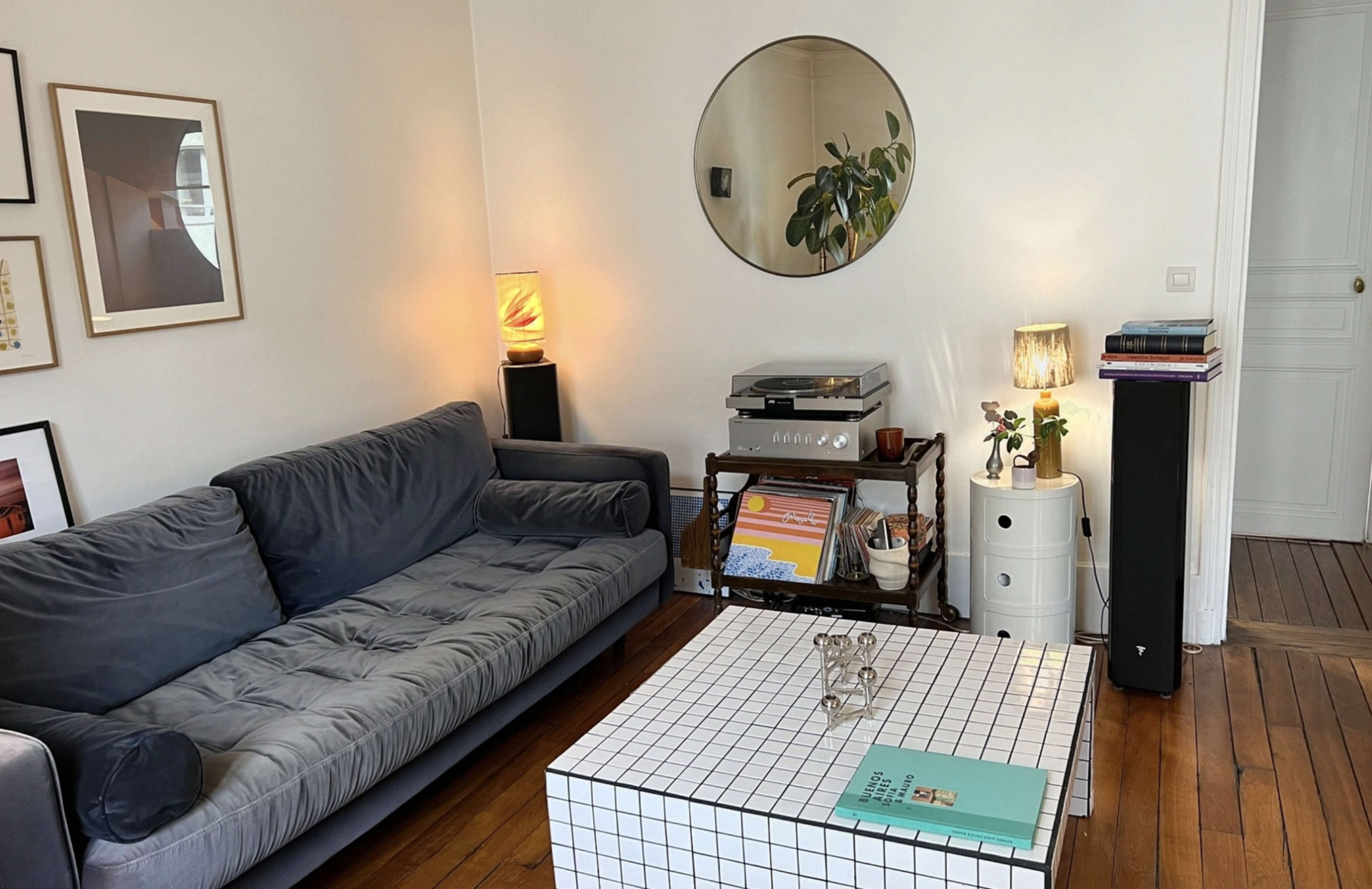 A modern living room features a gray sectional sofa, a round mirror, a coffee table with a tiled surface, a record player, and various decorative items.