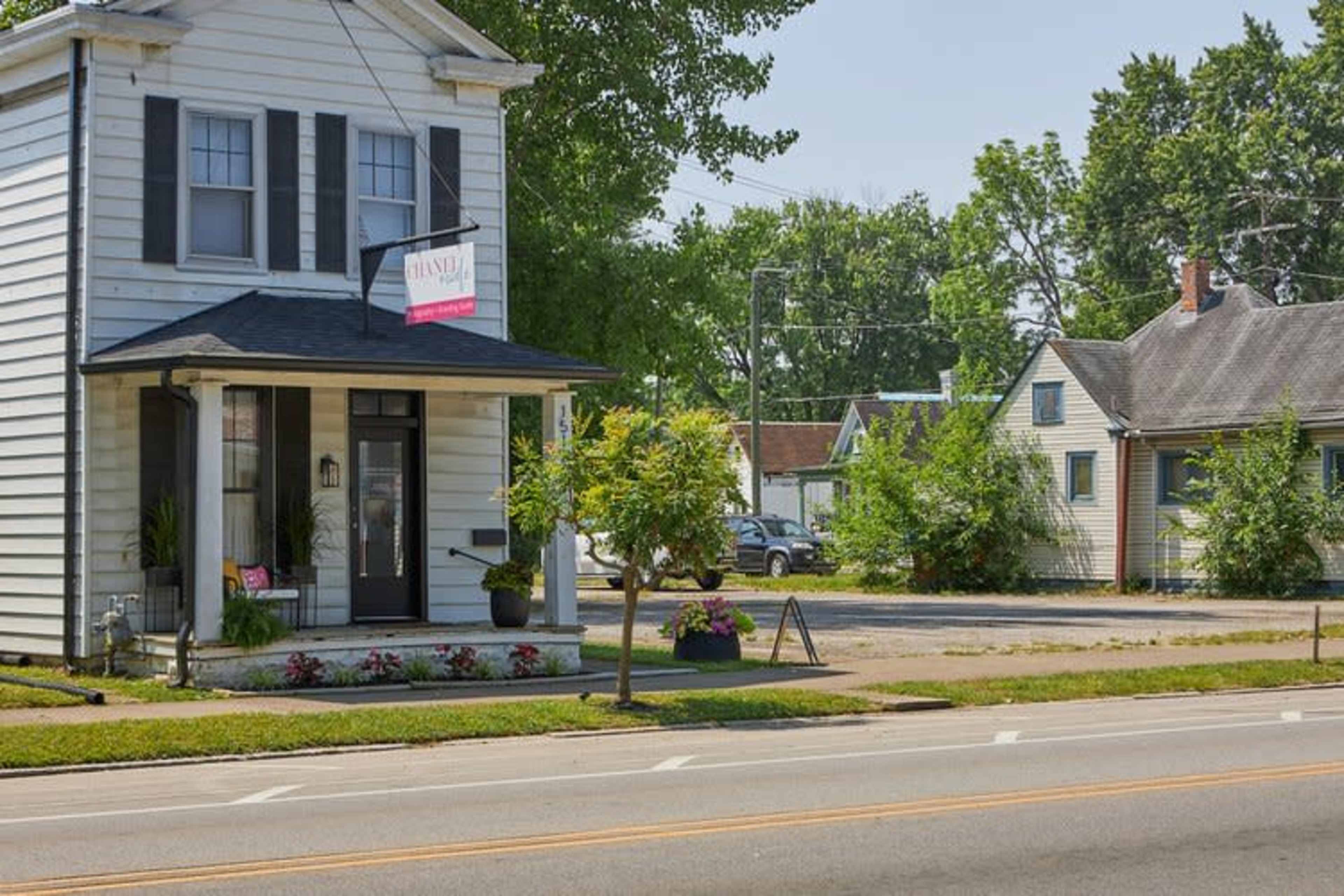 A small, two-story house with black shutters and a business sign on its porch stands next to a grassy area and several other buildings.