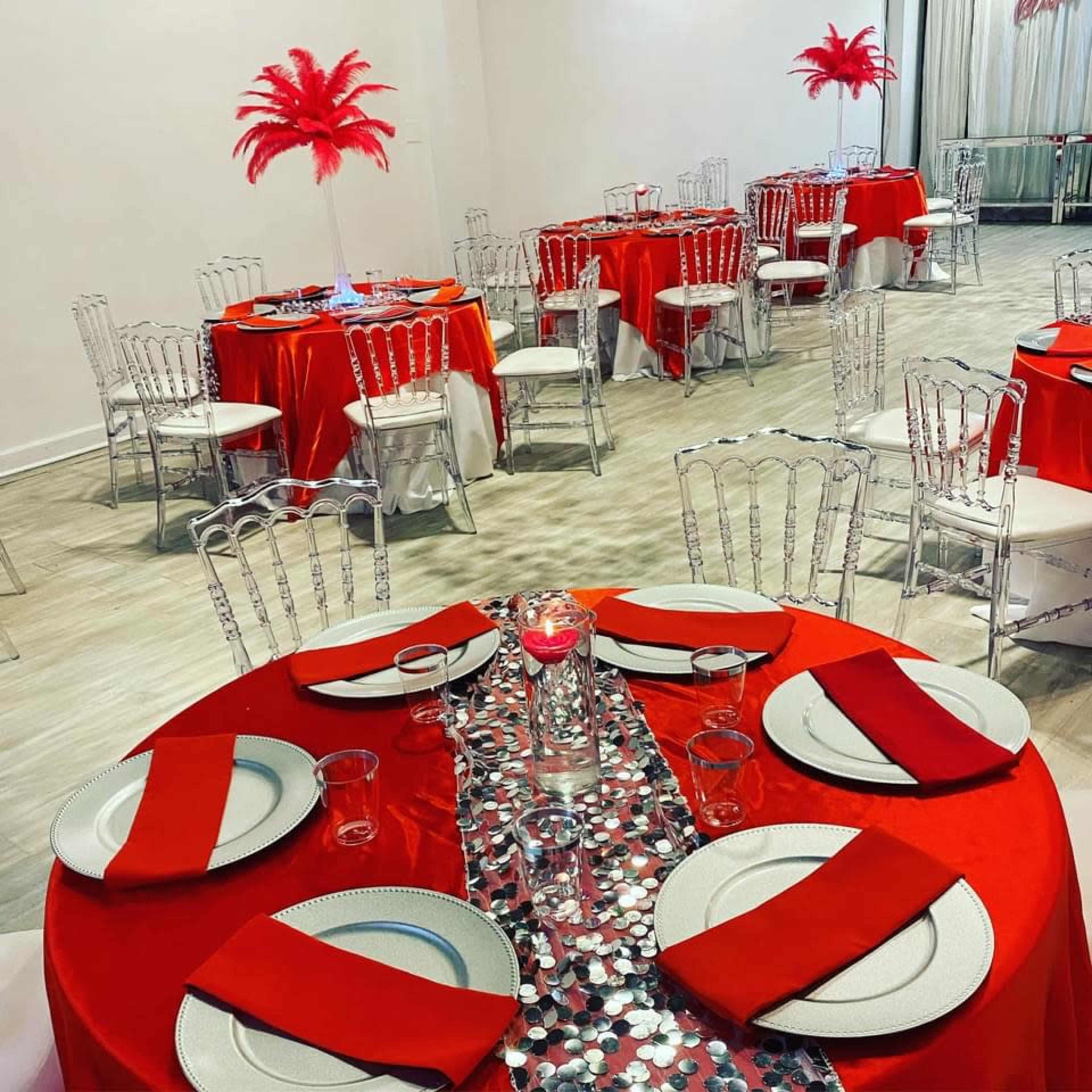 The image shows a banquet hall set for an event, featuring round tables covered in red cloth, white chairs, and centerpieces with red feather arrangements.