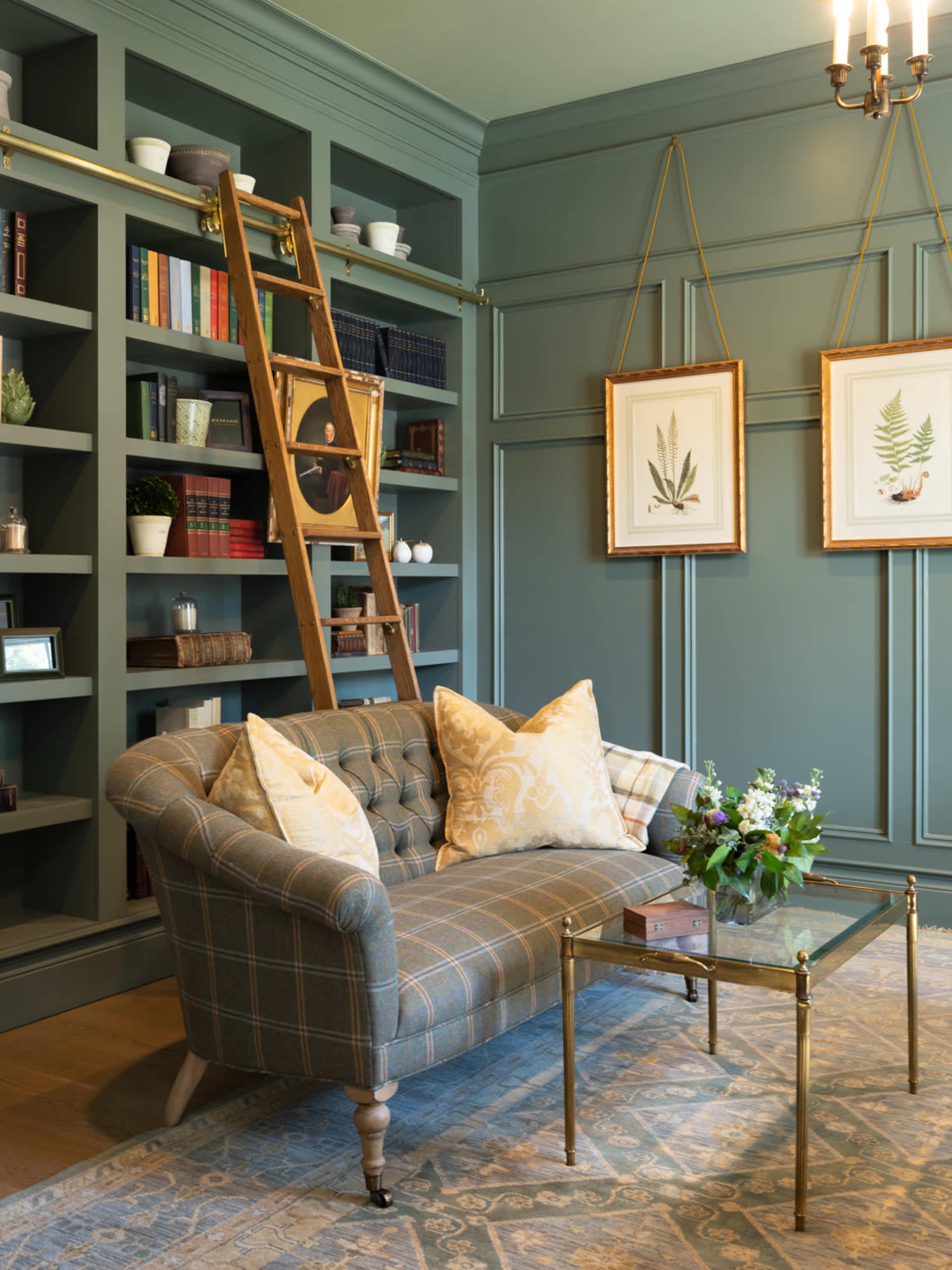 The scene features a cozy sitting area with a plaid sofa, a glass coffee table, and a wooden ladder against a dark green bookshelf filled with books and decorative items.