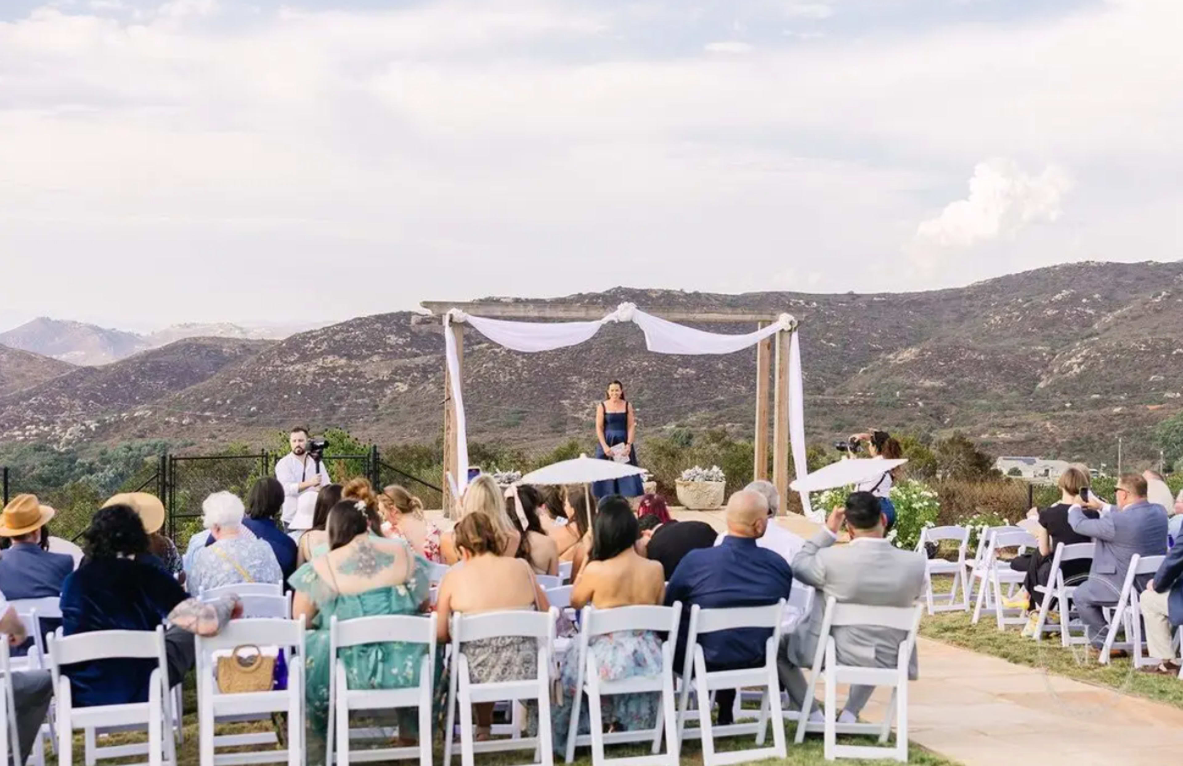 A wedding ceremony takes place outdoors with a decorated arch, surrounded by guests seated at tables and mountains in the background.