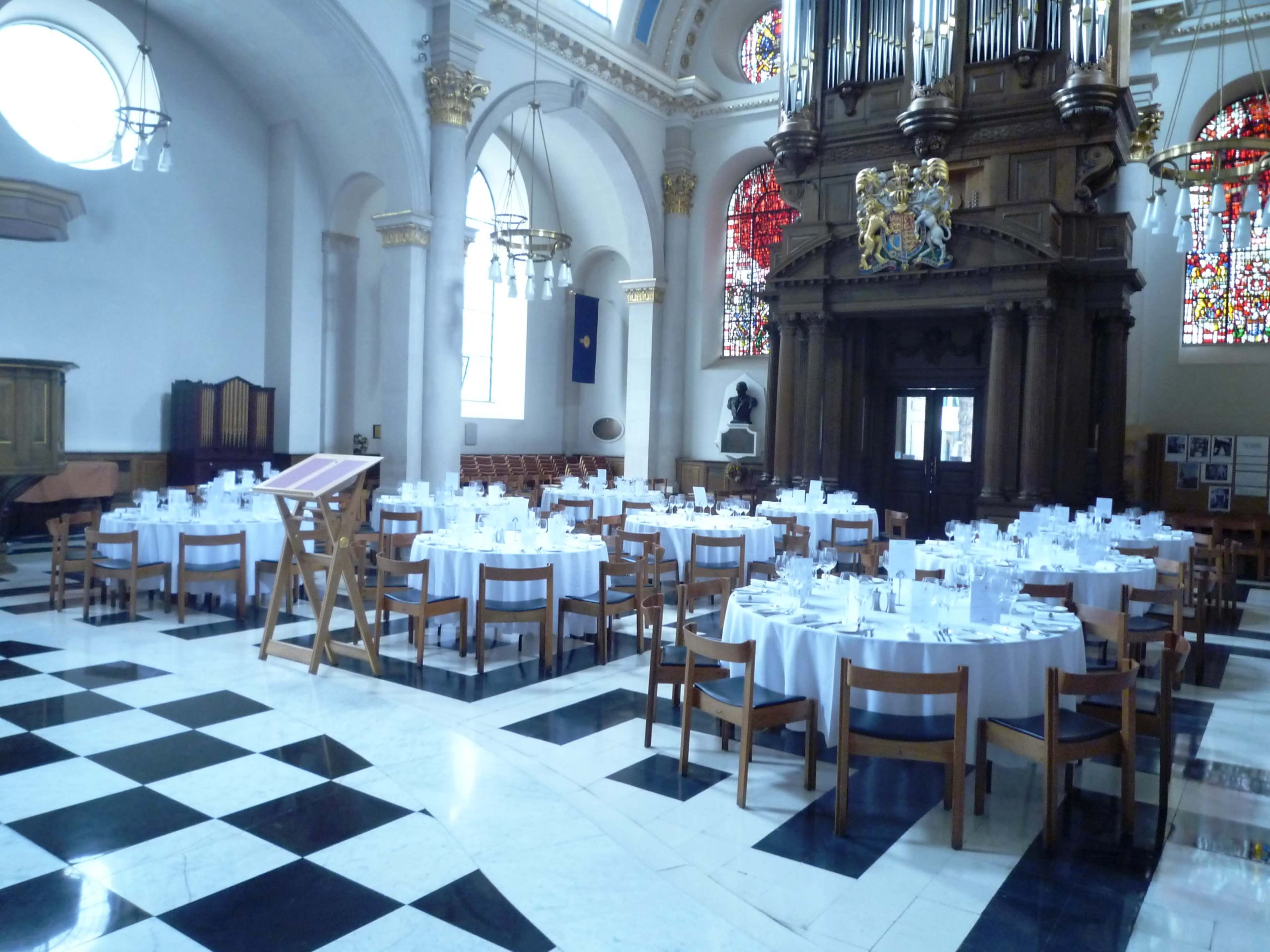 The image shows a large hall set up for a formal dining event, featuring several round tables covered with white tablecloths in front of a grand wooden organ and stained glass windows.