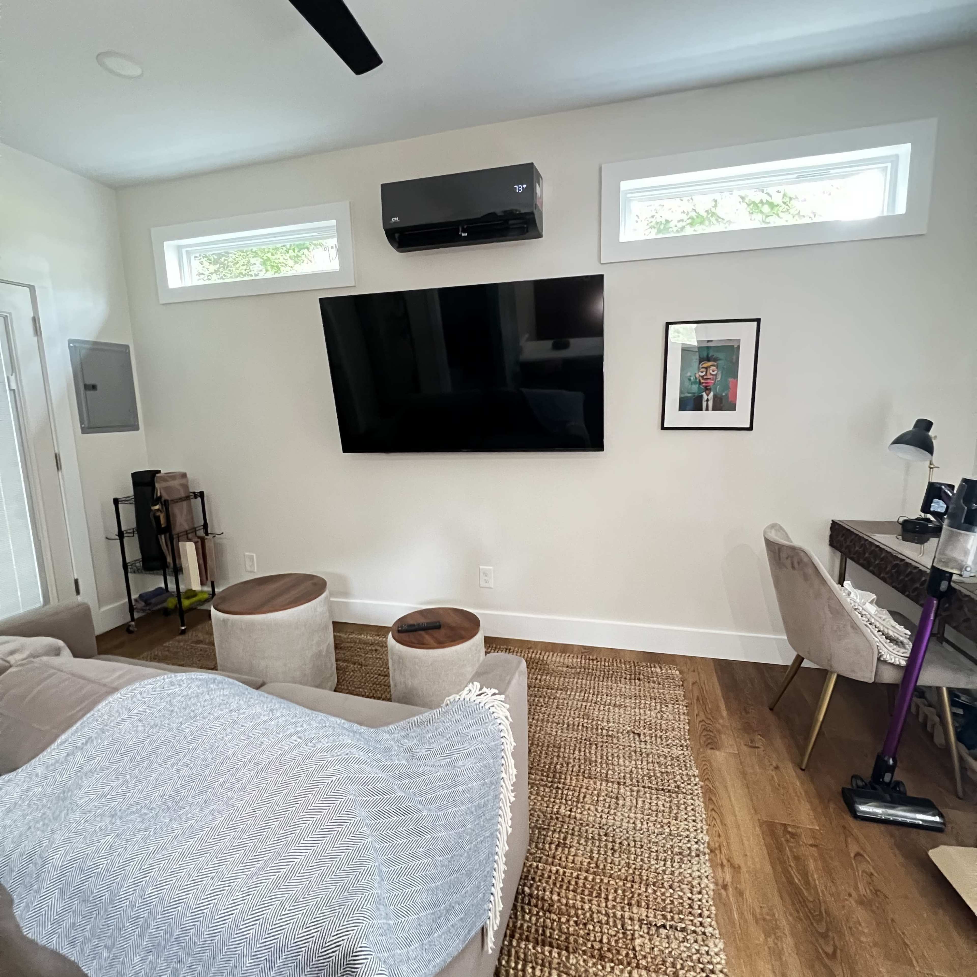 The image shows a modern living room with a wall-mounted television, a cozy sofa, two round stools, a small workspace with a desk and lamp, and large windows allowing natural light.