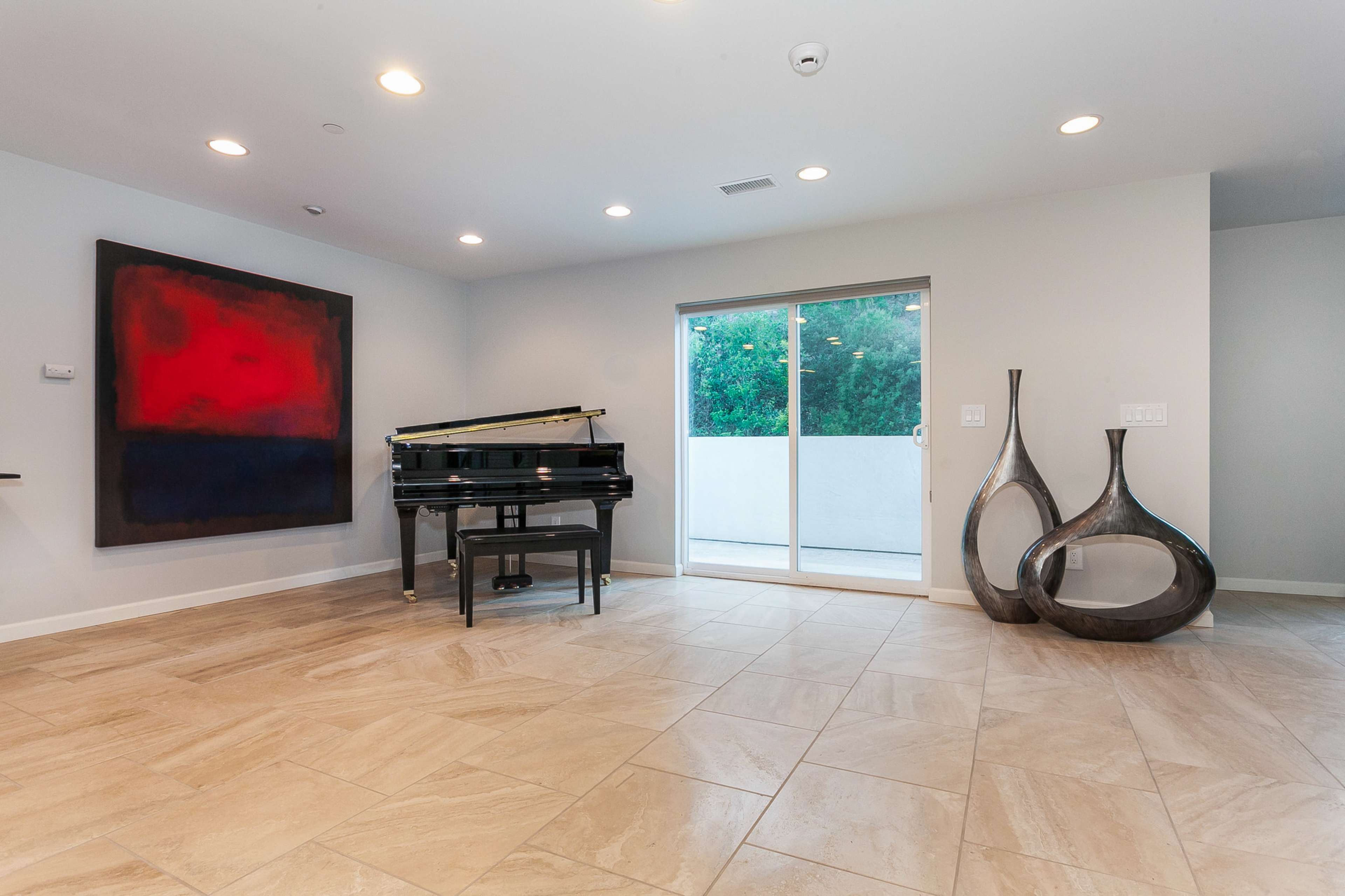 The image shows a spacious room featuring a black grand piano, a large red abstract painting, and two decorative vases near a sliding glass door leading to an outdoor area.