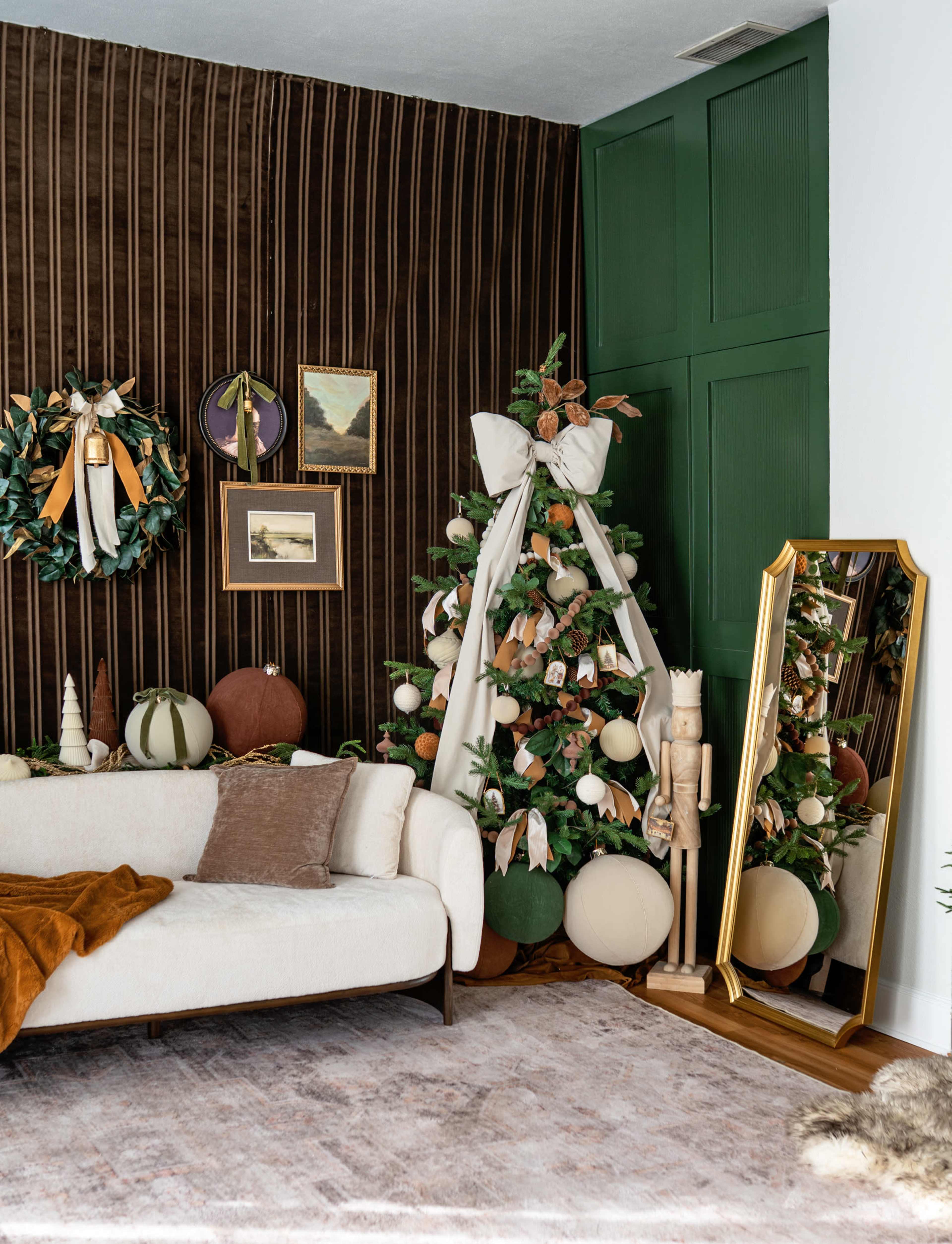 The image shows a living room corner decorated for the holidays, featuring a styled Christmas tree, wall decorations, a neutral-colored sofa, and a large mirror.