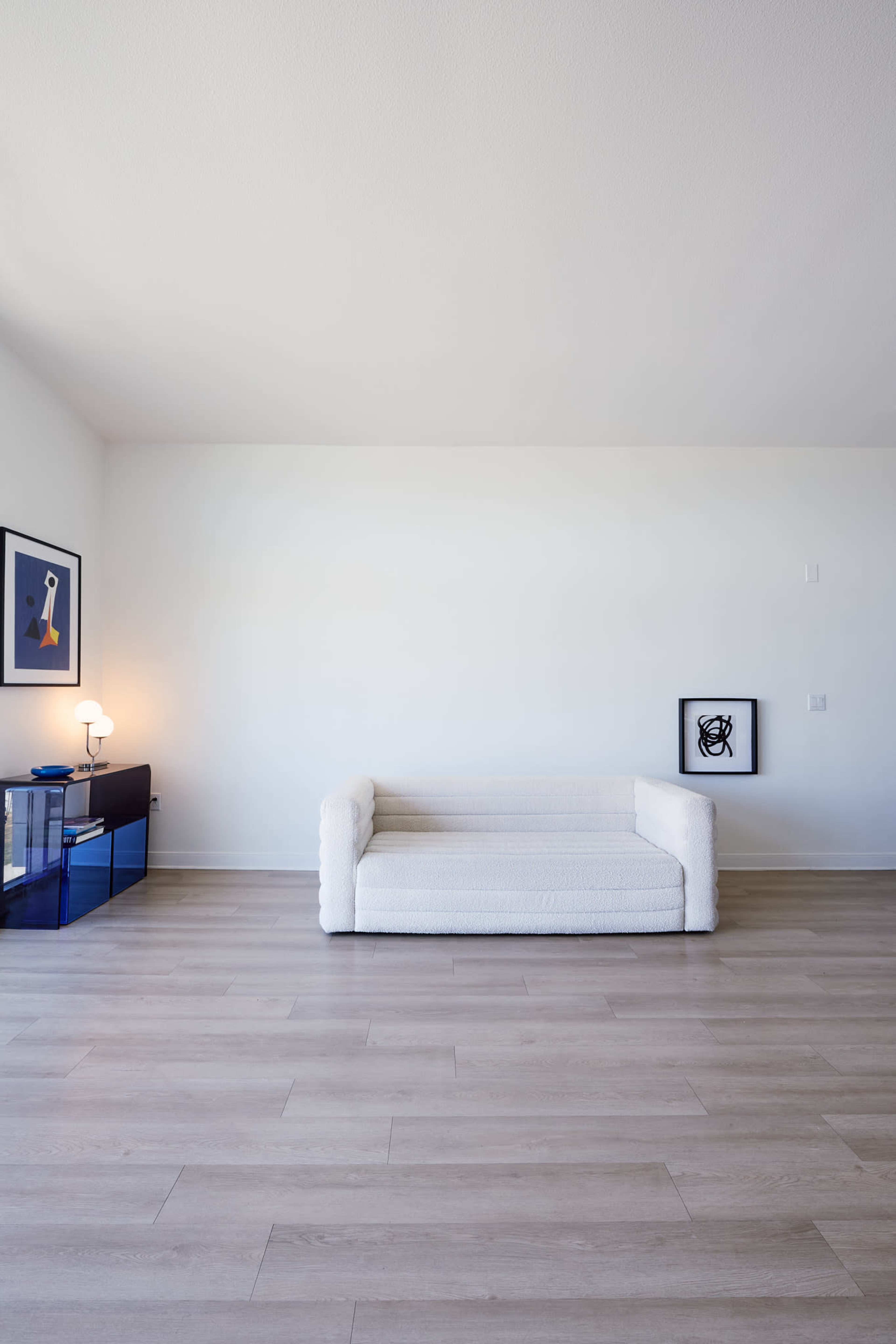 The image shows a minimalist living room with a white couch, a blue cabinet, and framed artwork on the walls.