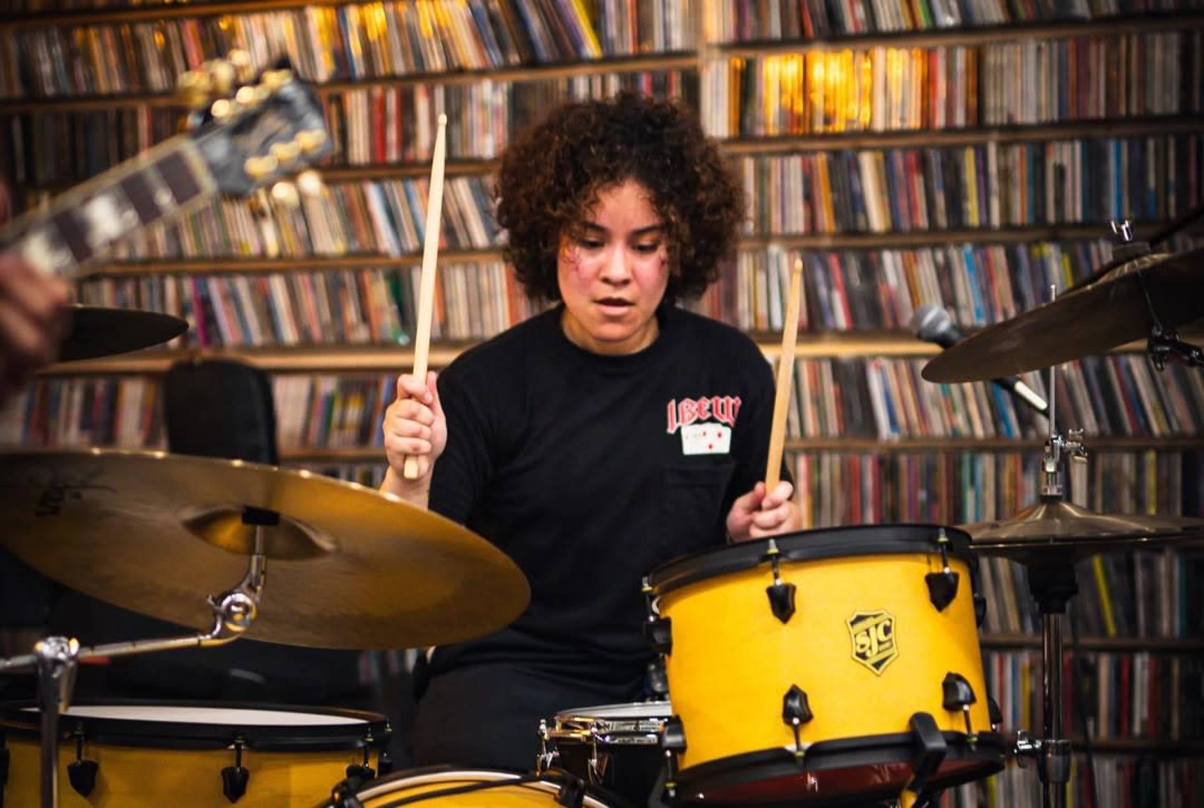 A drummer with curly hair plays a yellow drum set in front of a wall filled with CDs.