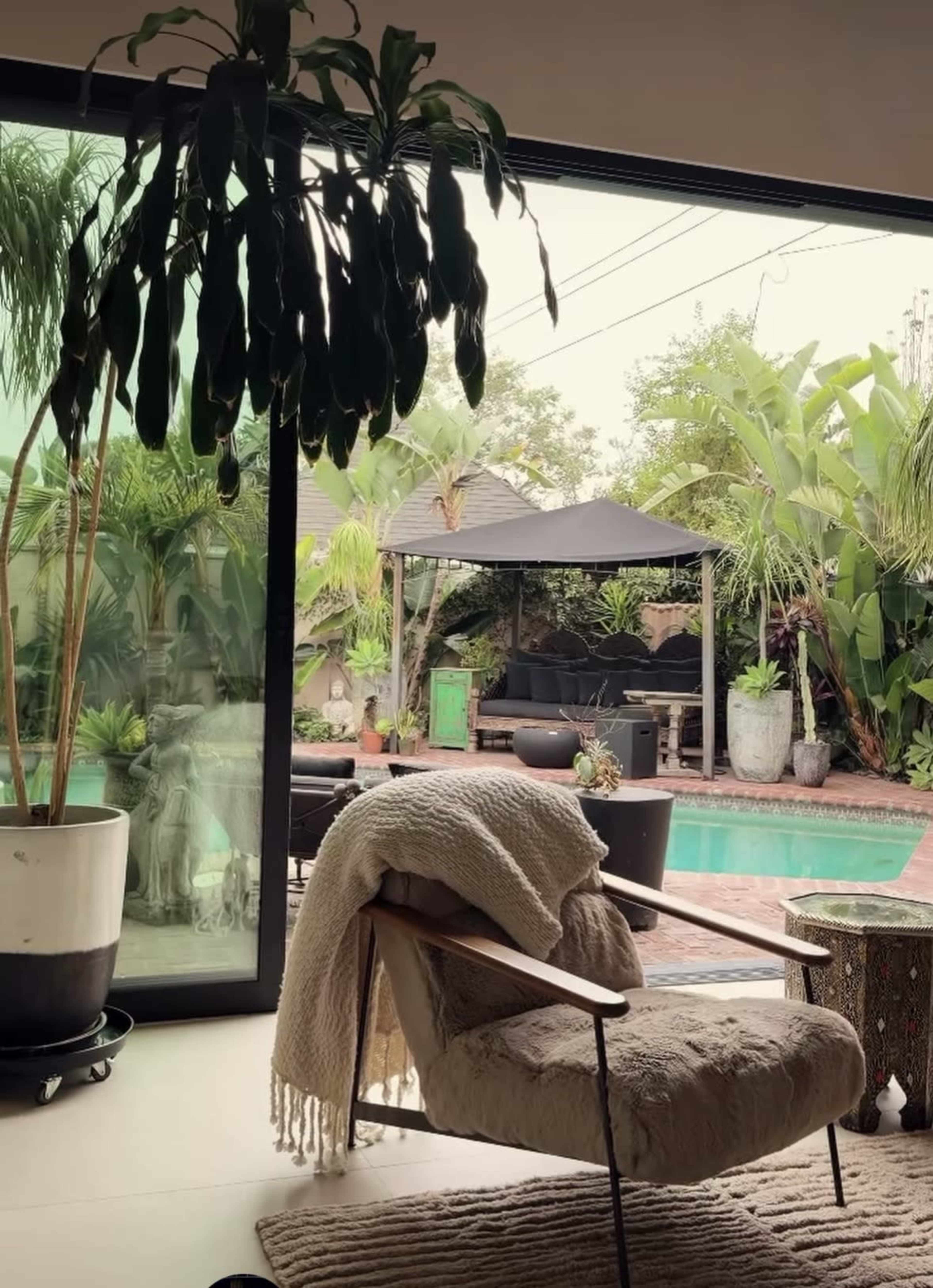 A modern indoor space features a cozy chair with a blanket, overlooking a lush outdoor area with a pool and a shaded seating arrangement surrounded by tropical plants.