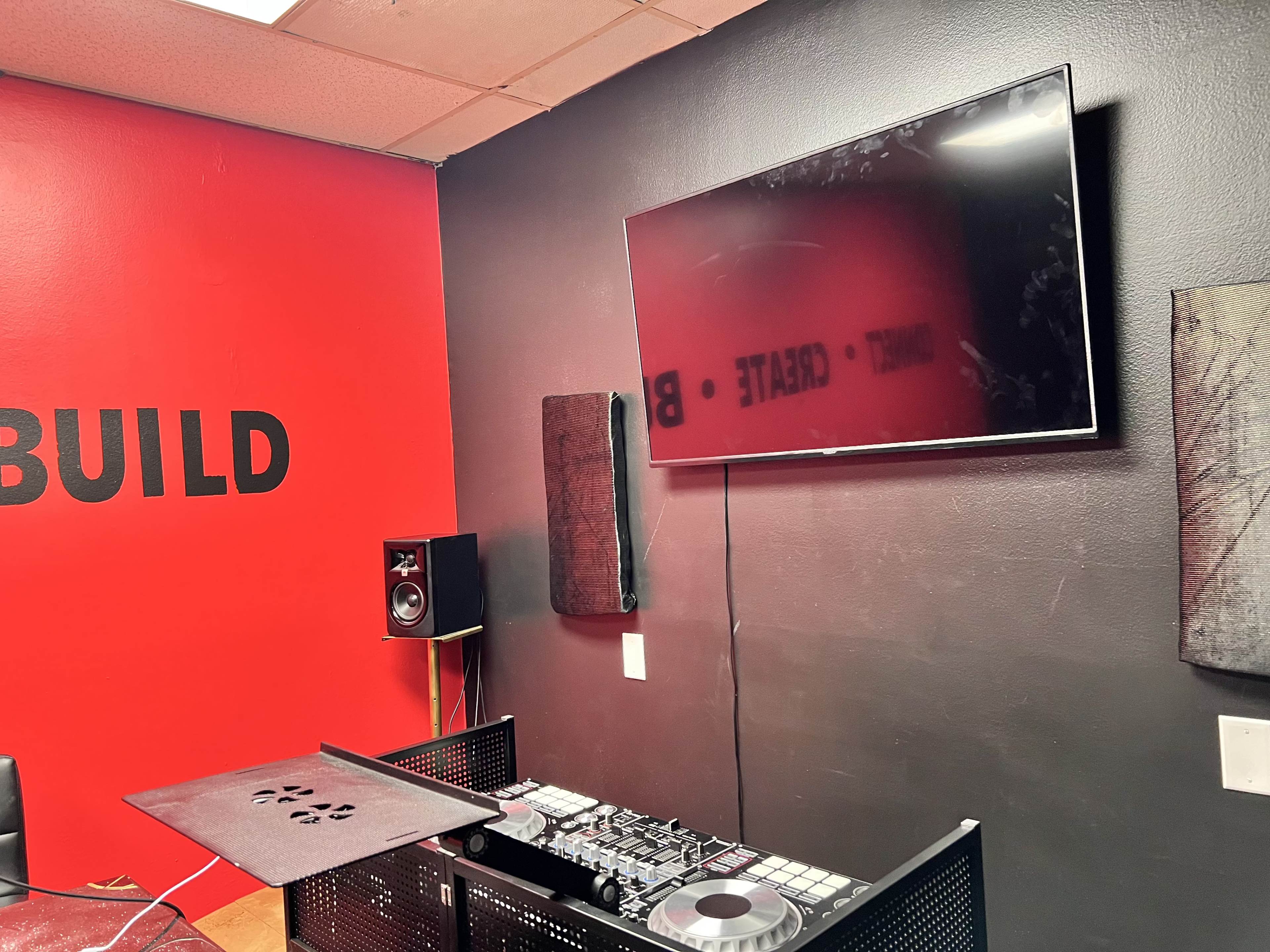 A room with a red and black color scheme featuring a television mounted on the wall, a sound speaker, and a DJ equipment setup on a table.
