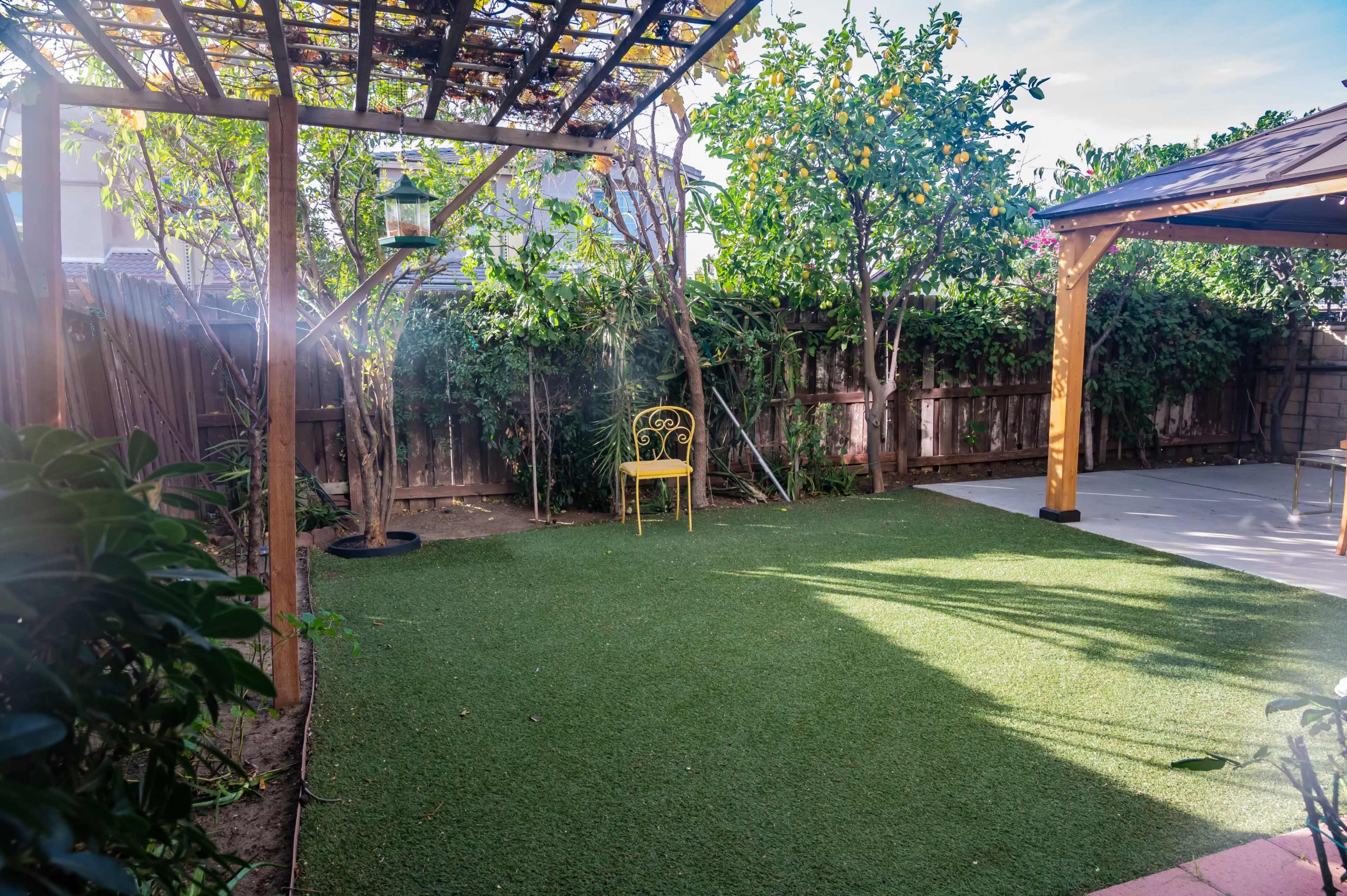 The scene shows a backyard with artificial grass, a solitary yellow chair under a pergola, and lush greenery surrounding the area.