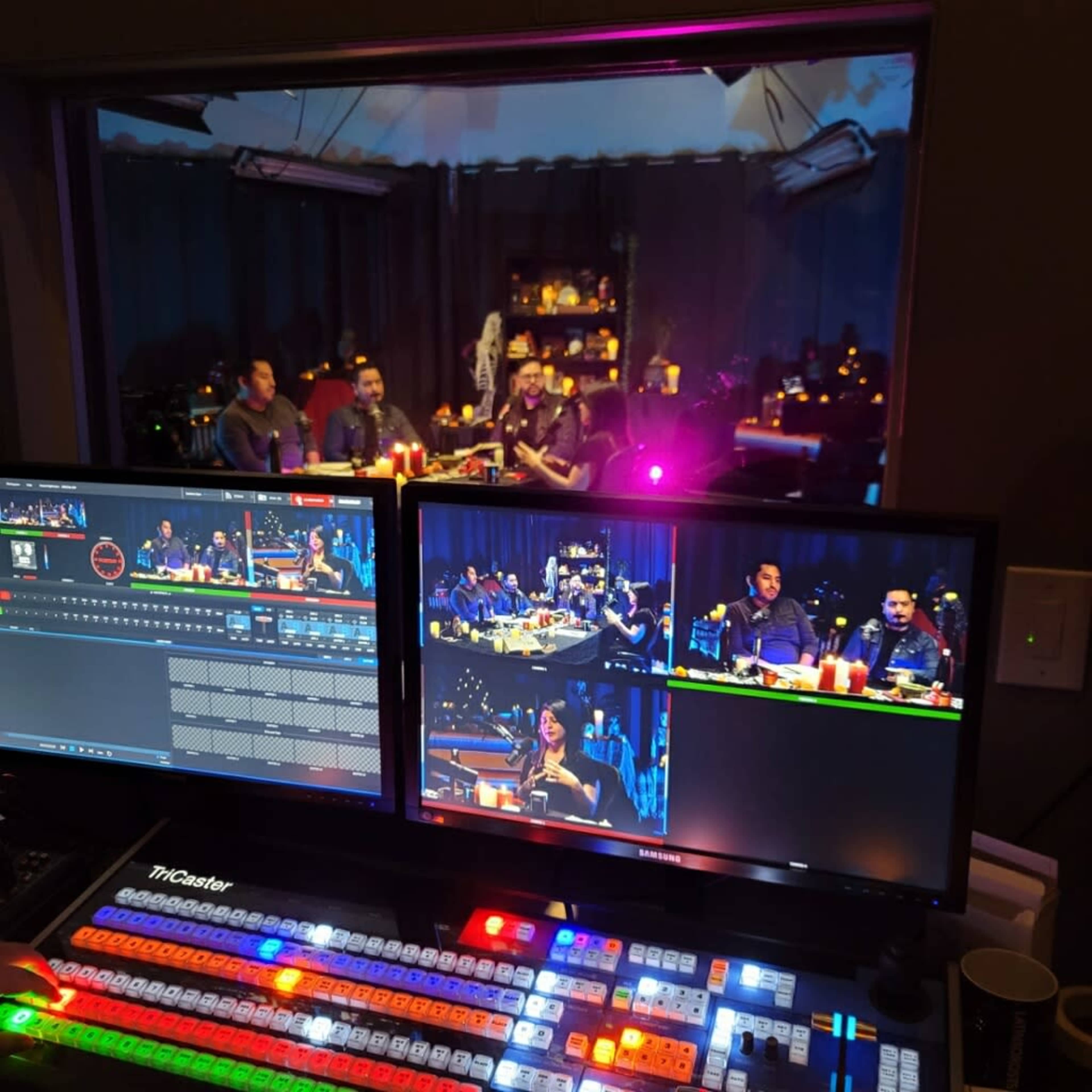A broadcast control room with two monitors displaying a live panel discussion and a window showing the panelists seated at a table with equipment in front.