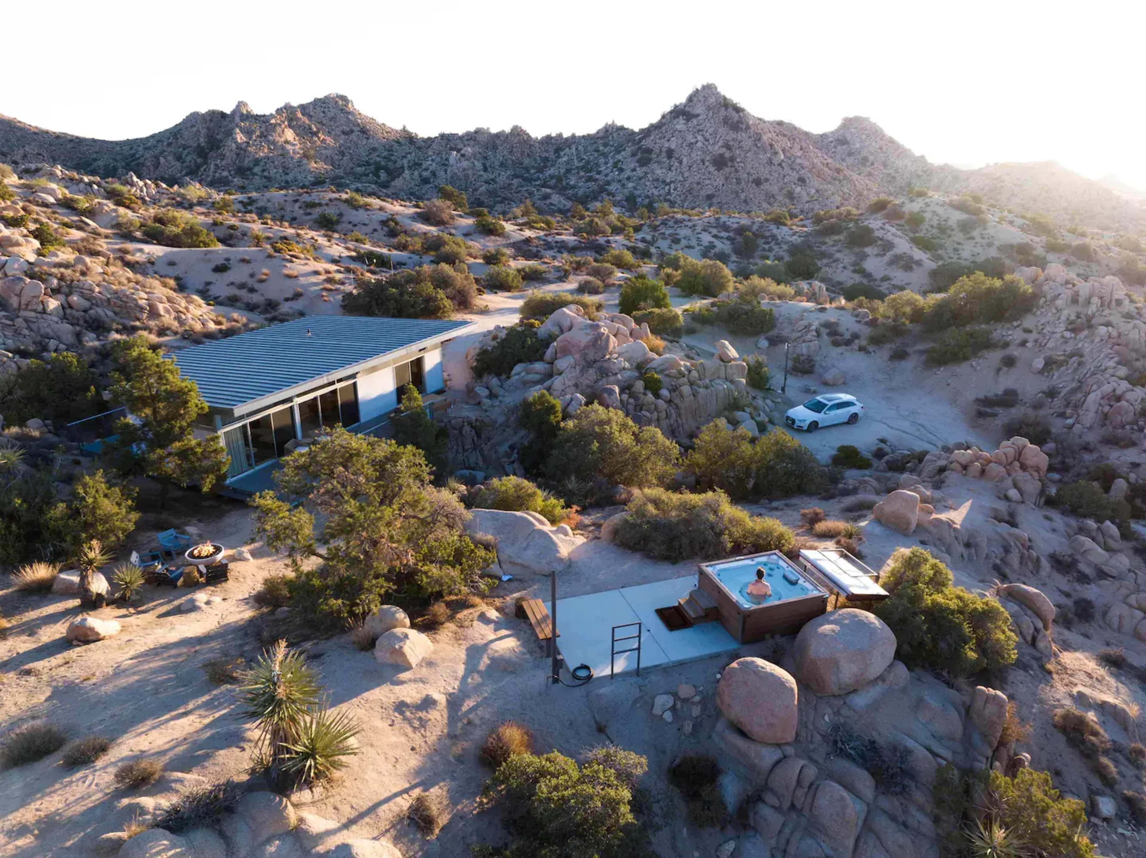 A modern house with a hot tub is situated among large rocks in a desert landscape with mountains in the background.