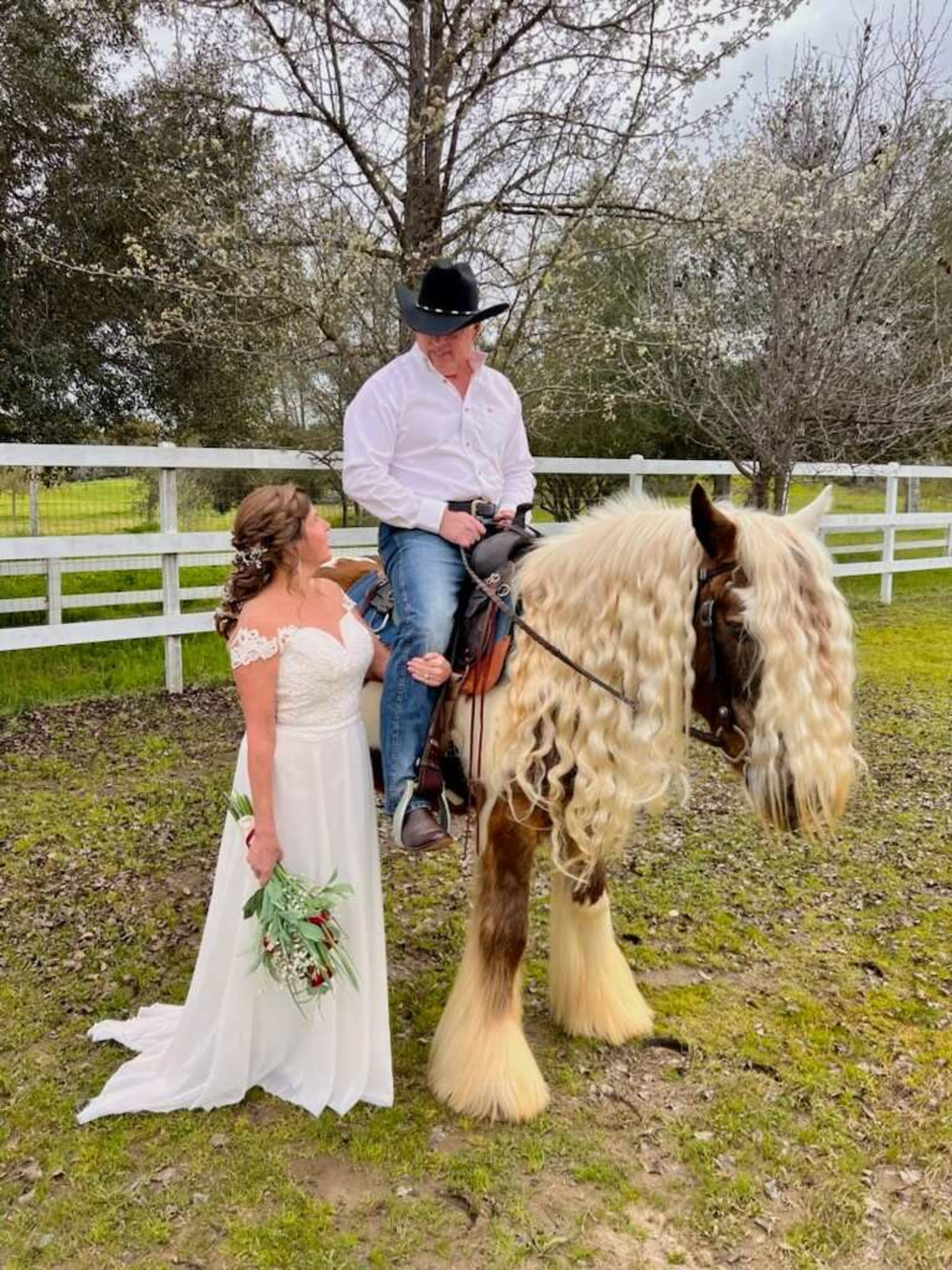 A woman in a white wedding dress stands beside a man in a cowboy hat seated on a horse with long, flowing hair.