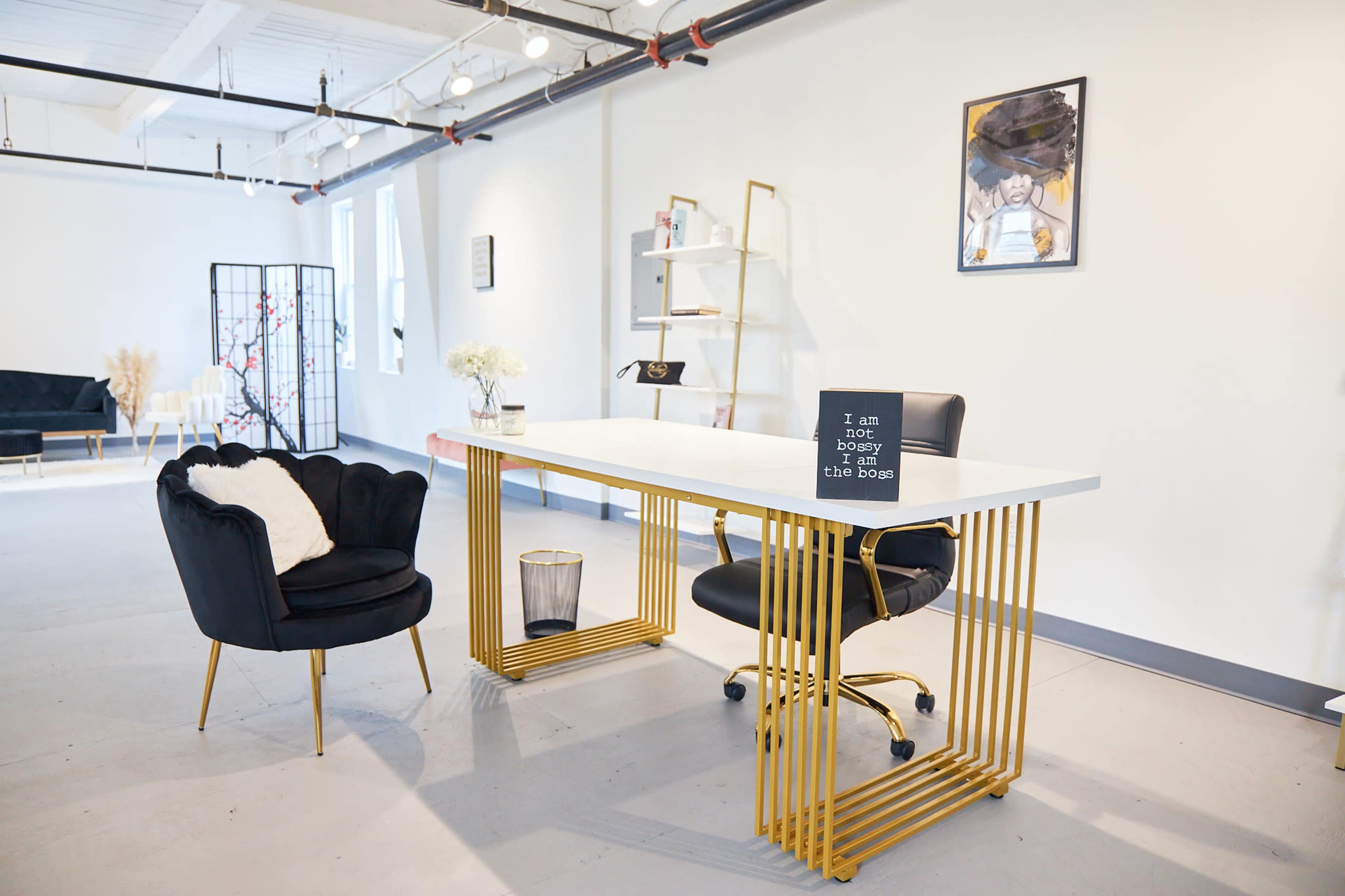 A modern office space features a sleek white desk with gold metal legs, a black chair, and a decorative sign, accompanied by a plush armchair and minimalist shelving in the background.