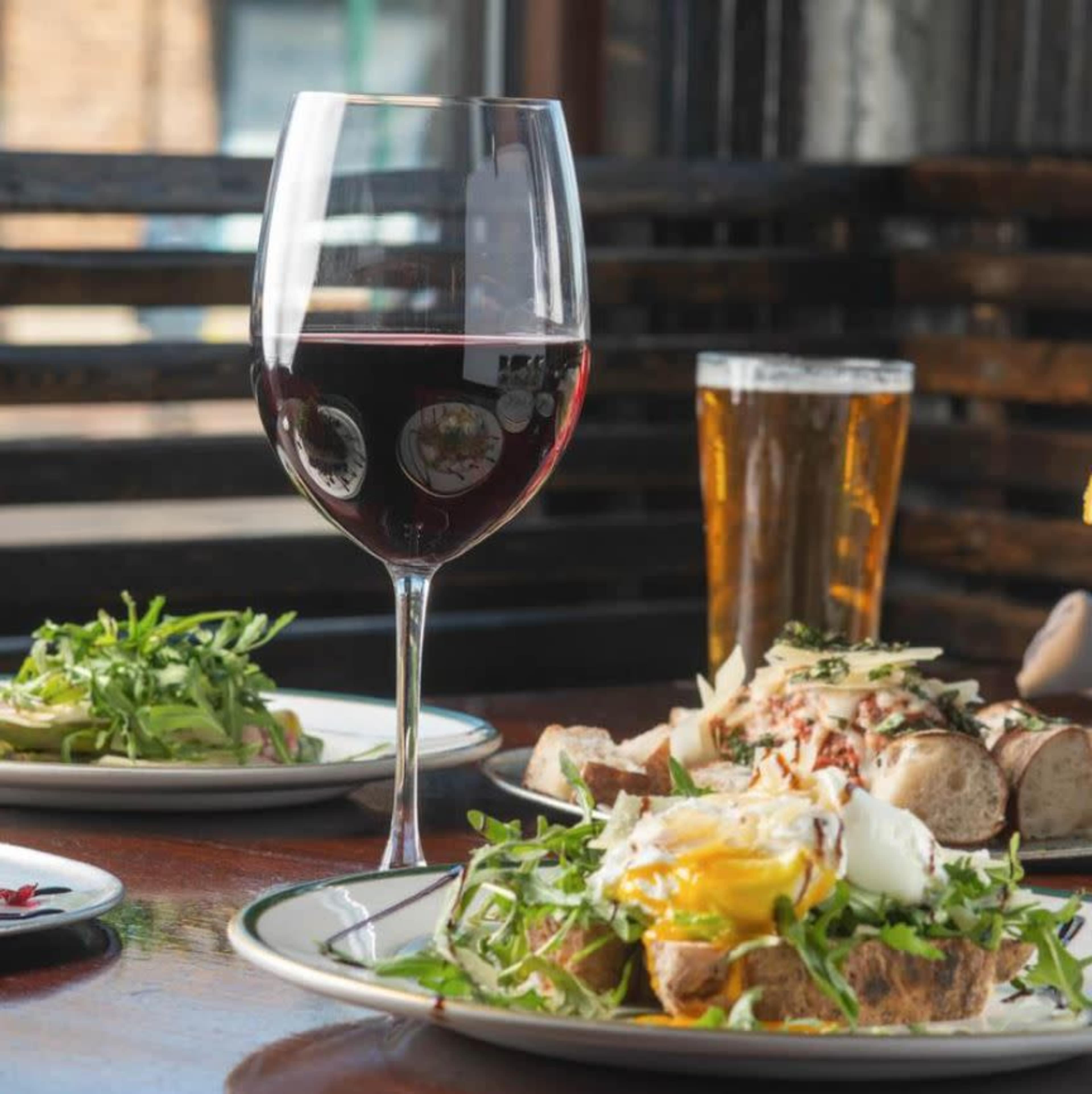 A glass of red wine sits beside a plate of salad, a dish with bread and topped egg, and a pint of beer on a wooden table in a restaurant setting.