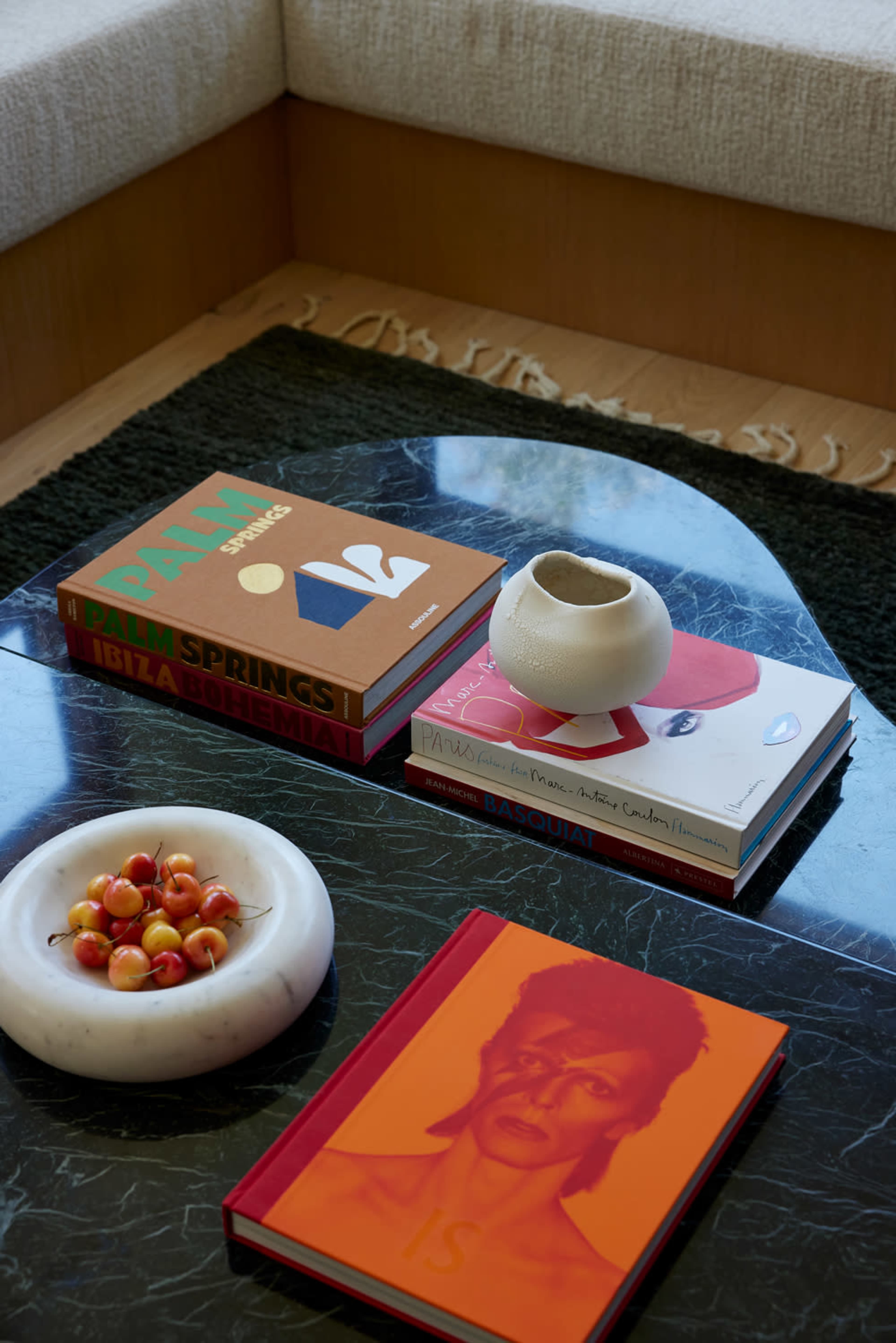 A coffee table features several art and design books stacked on top of a dark marble surface, accompanied by a small ceramic vase and a bowl of cherries.