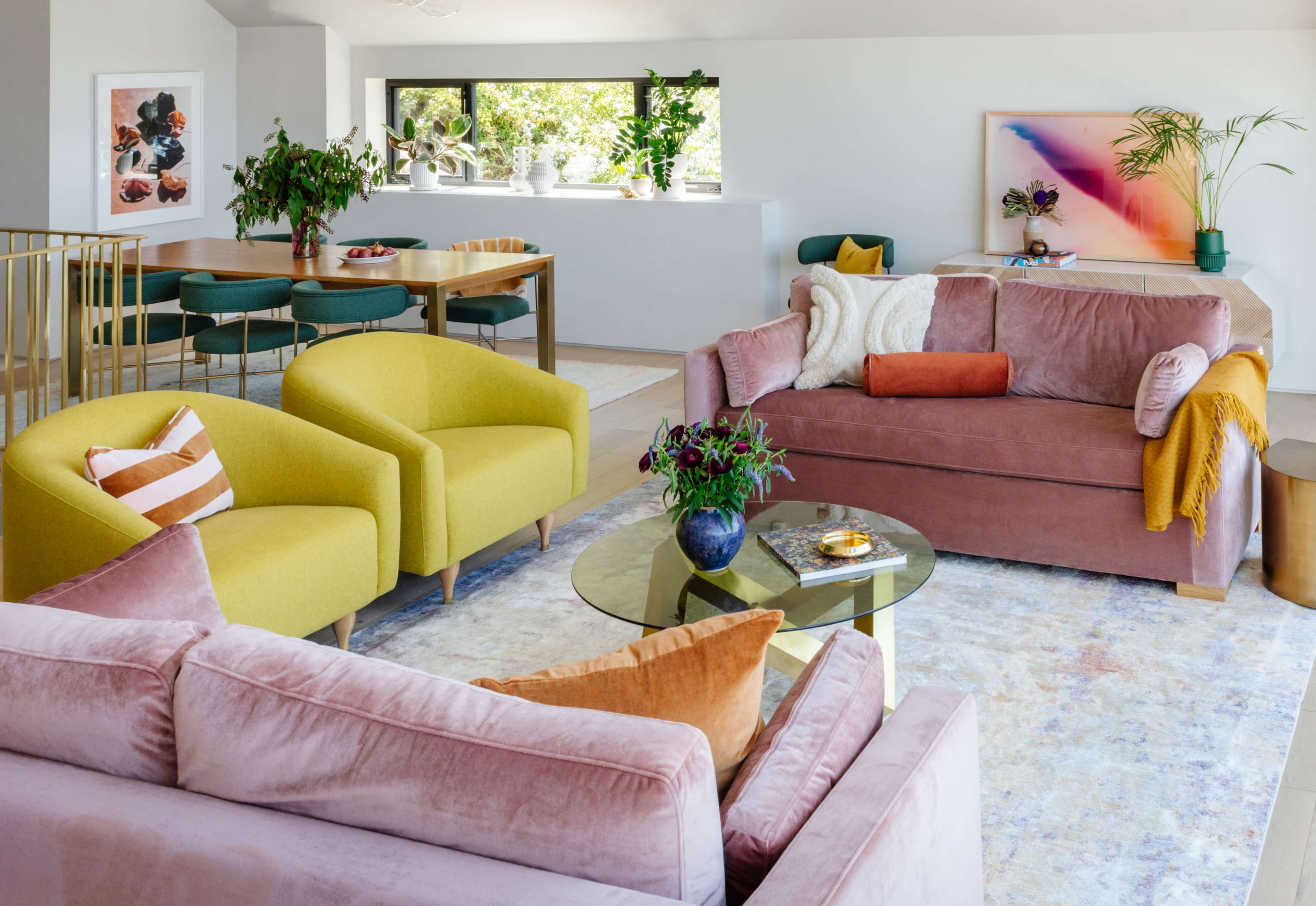A modern living room featuring two plush sofas, bright yellow armchairs, a glass coffee table, and a dining area with green chairs, all surrounded by plants and artwork.