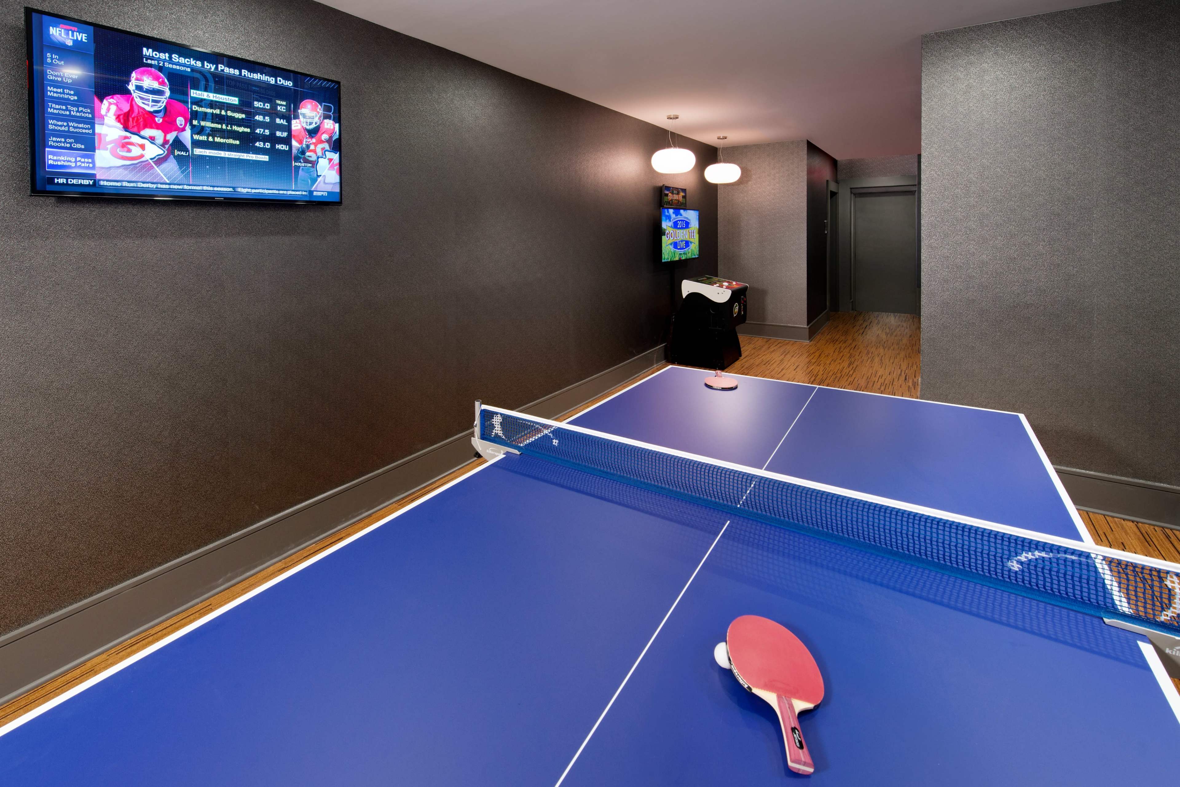 A blue ping pong table is set up in a game room with a television displaying sports statistics on one wall.