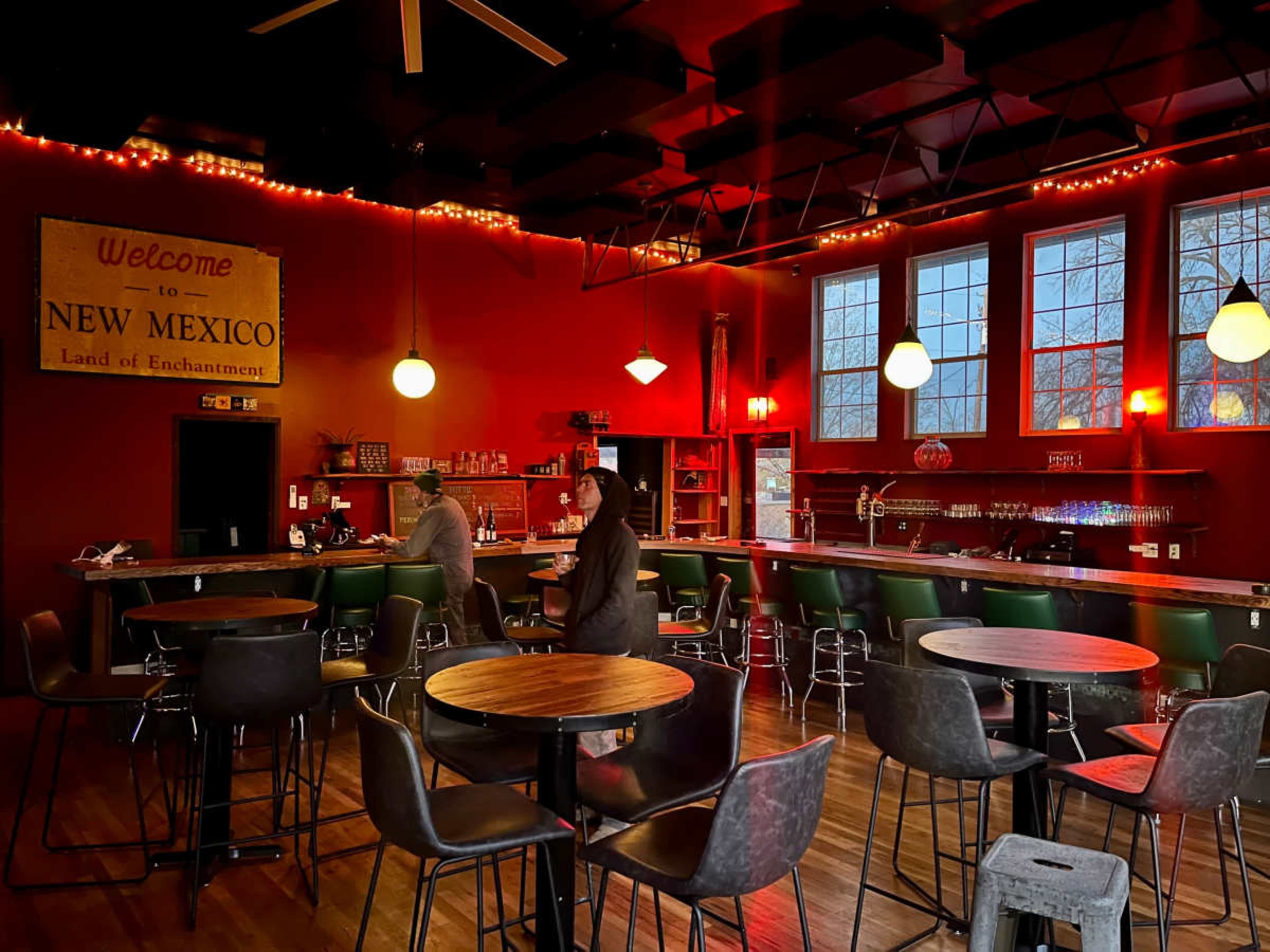 The image shows a dimly lit bar with red walls, several tables and chairs, and large windows letting in natural light.