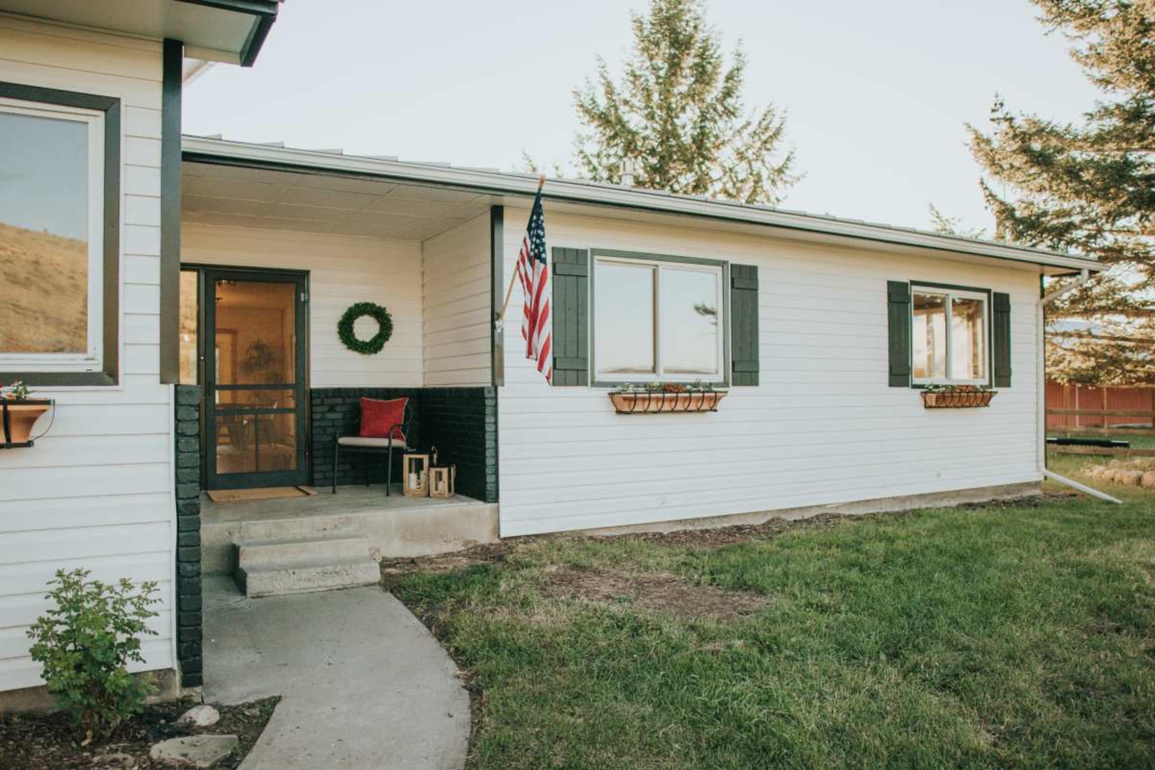 A white single-story house with green shutters has a porch featuring a small seating area and an American flag hanging by the door.