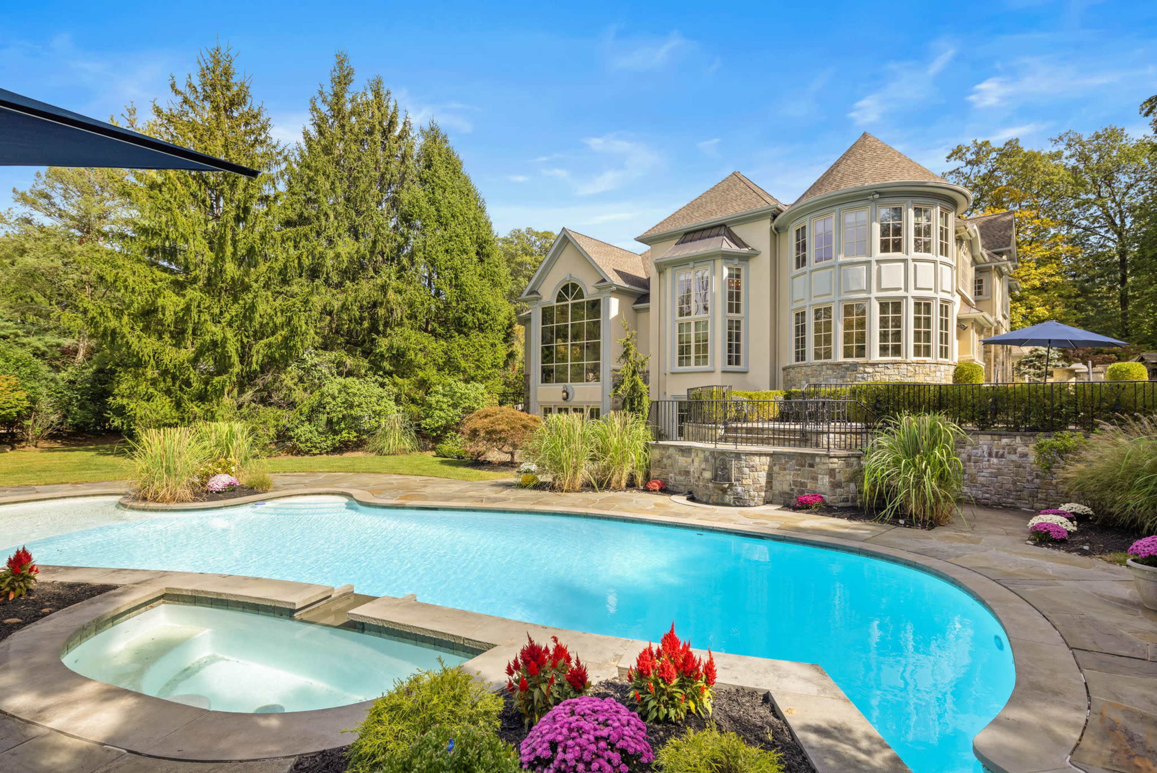 The image features a large swimming pool surrounded by a well-maintained garden and a modern house with multiple windows and a distinctive architectural design.