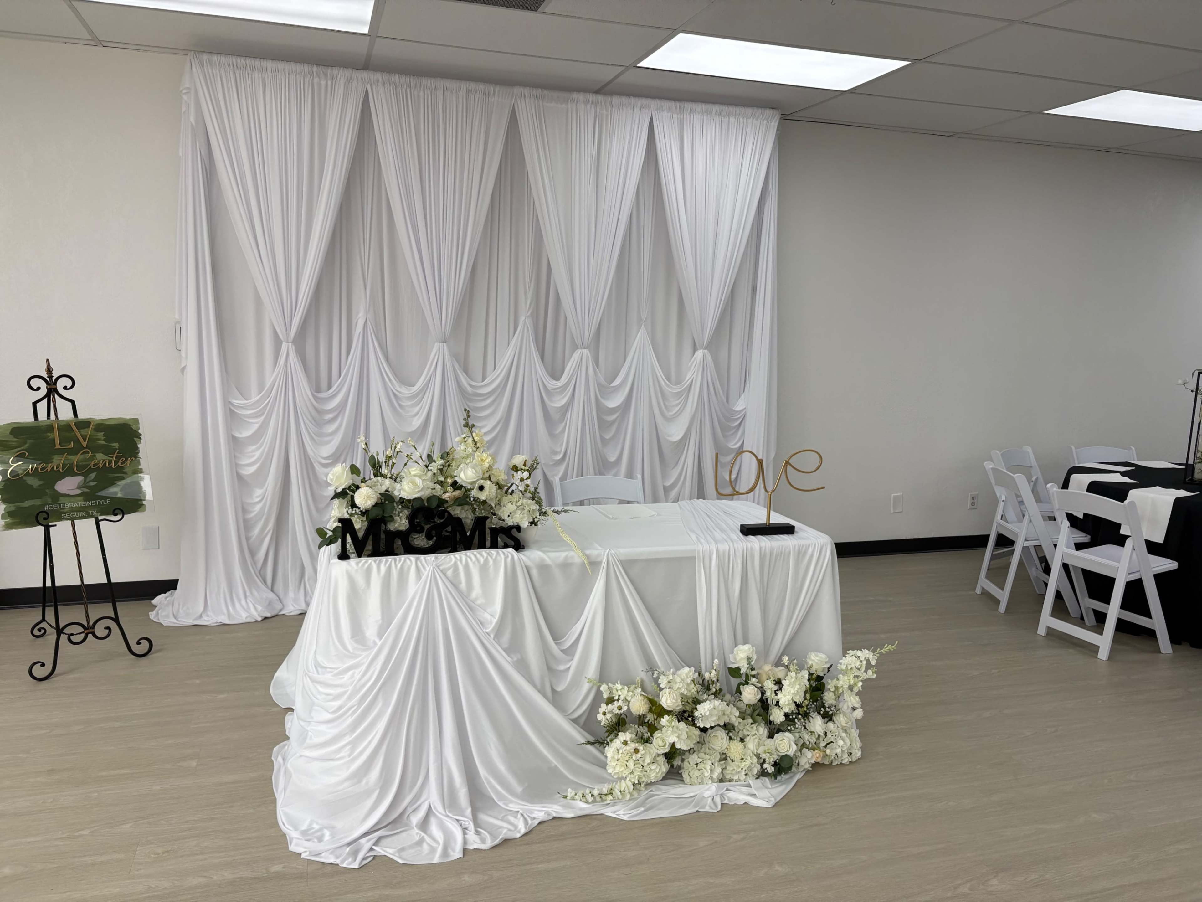 The image shows a decorated wedding reception area featuring a white draped backdrop, a head table adorned with flowers, and a "Mr. & Mrs." sign along with a decorative "love" piece.