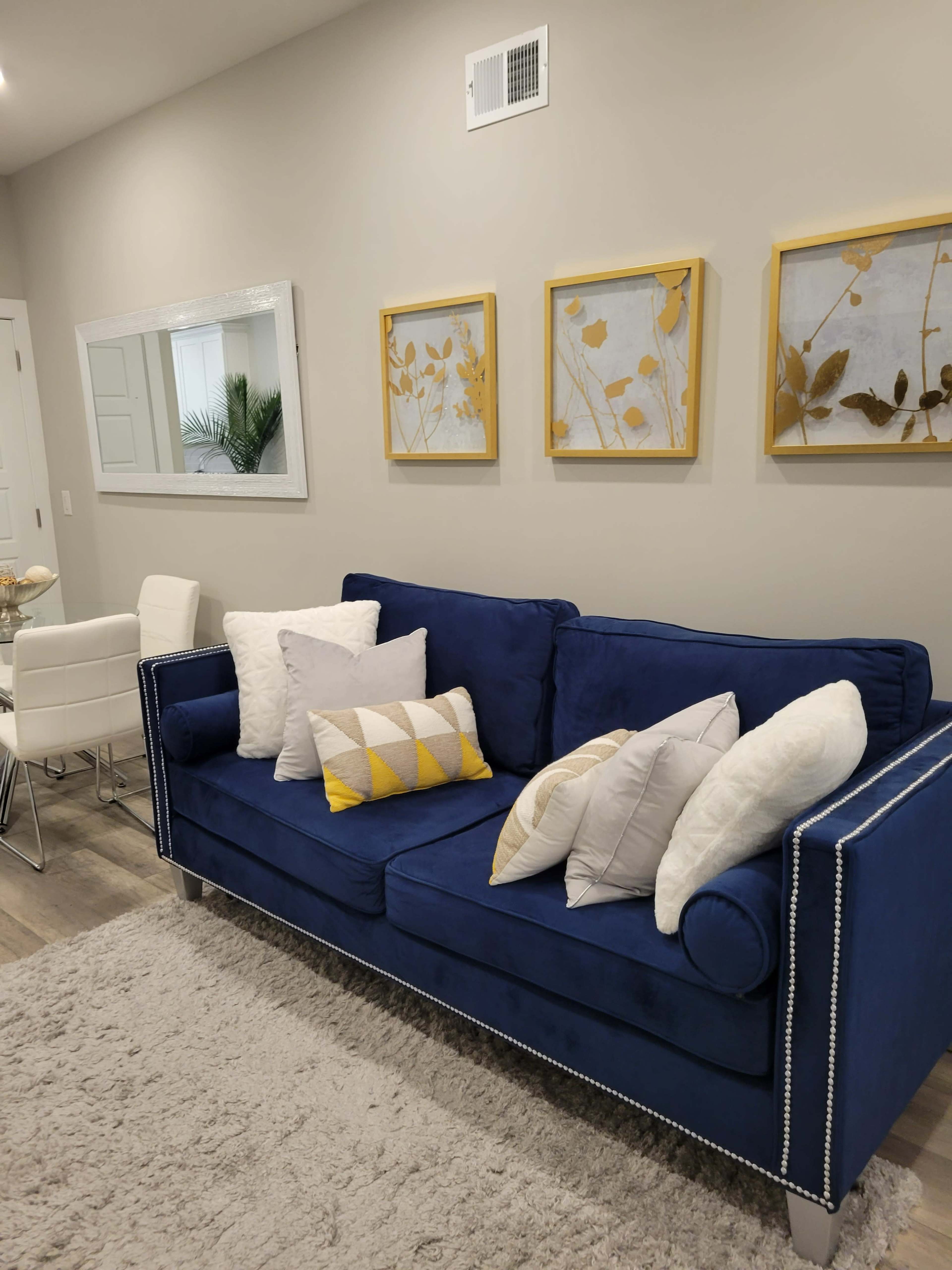 A blue couch with various patterned pillows is positioned against a wall adorned with gold-framed art pieces, beside a dining area with white chairs and a table.