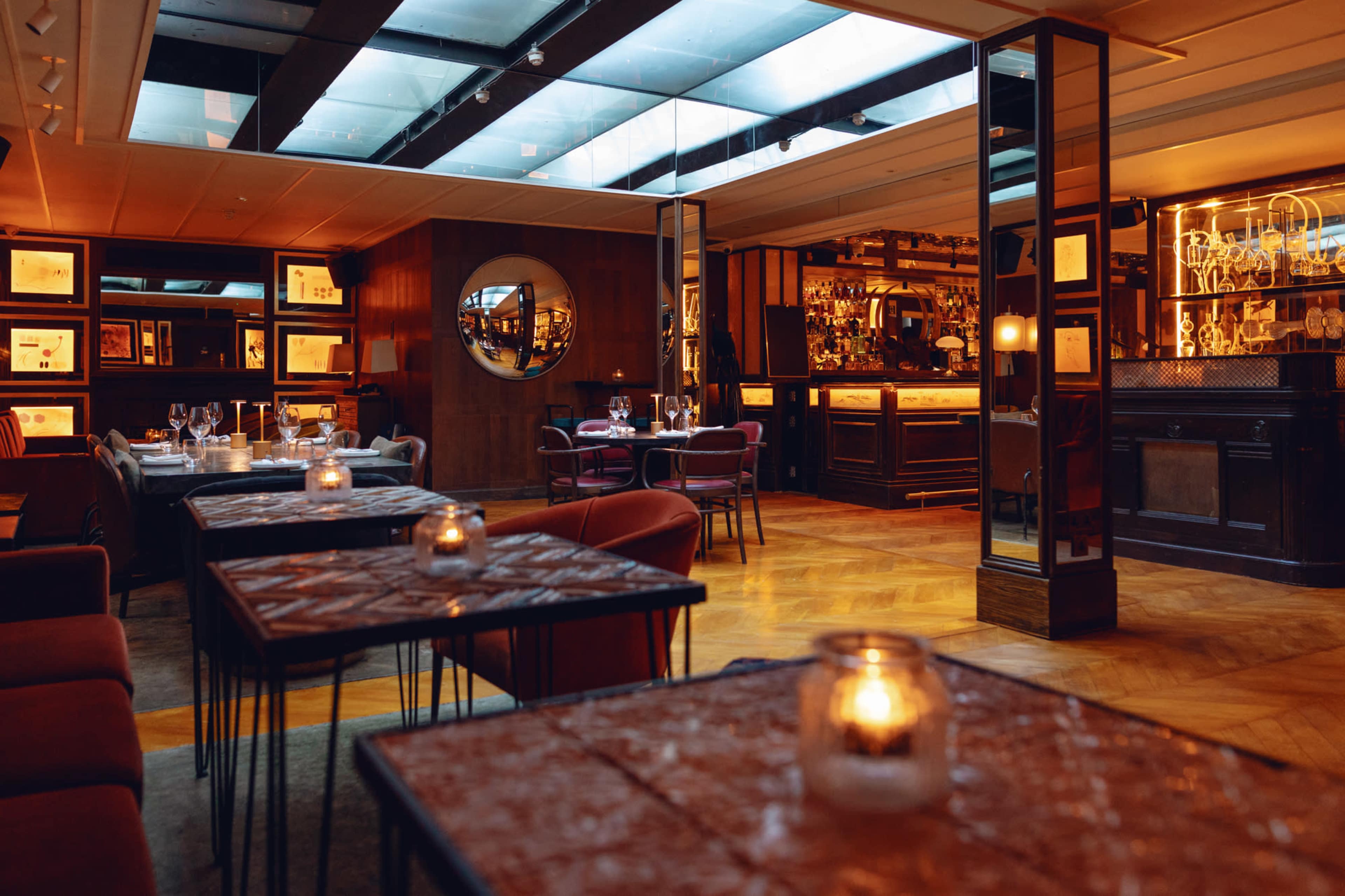 An elegant restaurant interior with wooden accents, tables set for dining, and a bar in the background illuminated by warm lighting.
