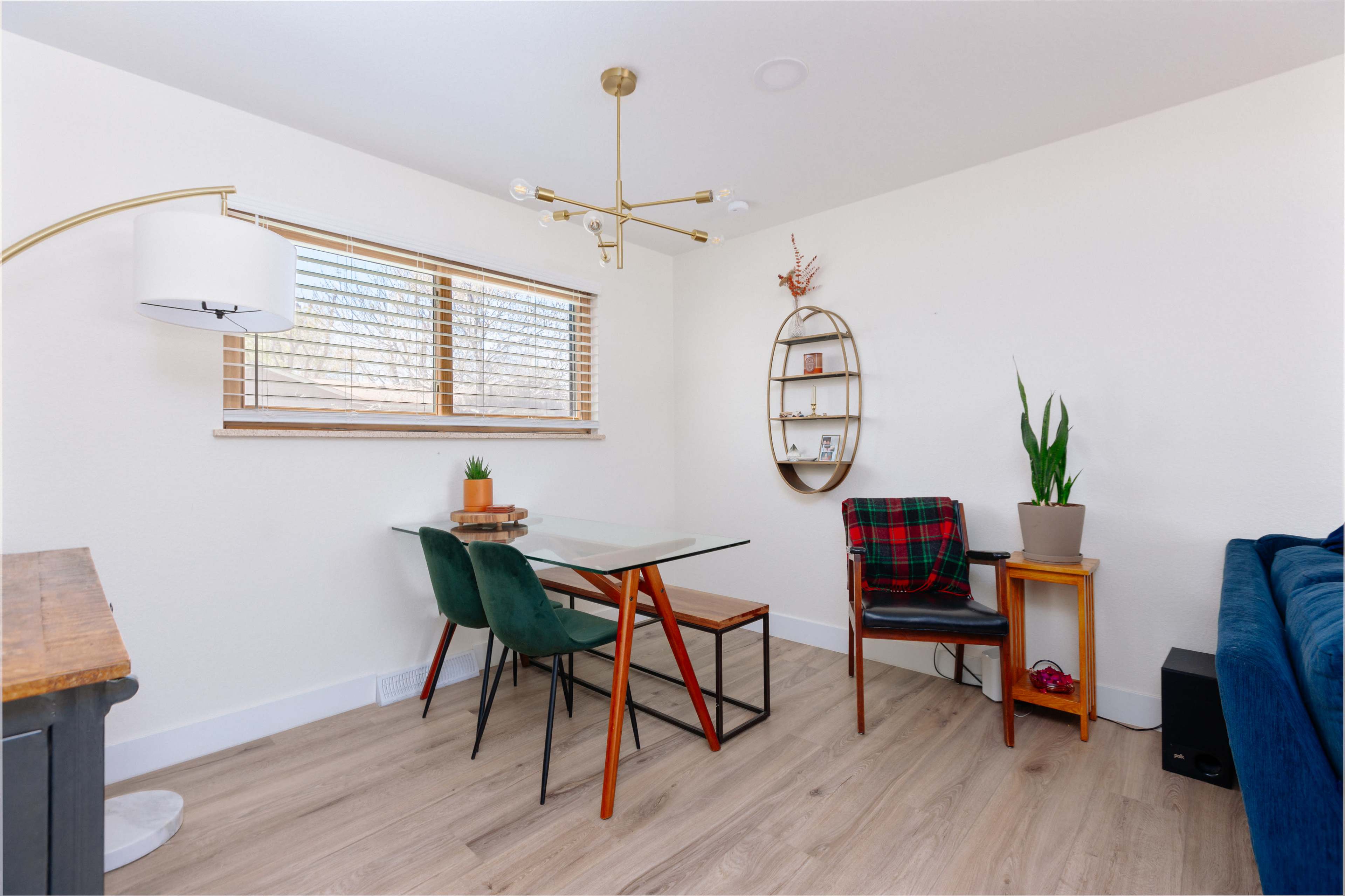 A small dining area featuring a glass table with wooden legs, two green chairs, a single wooden chair, a floor lamp, a wall-mounted shelf, a potted plant, and a sofa in the background.
