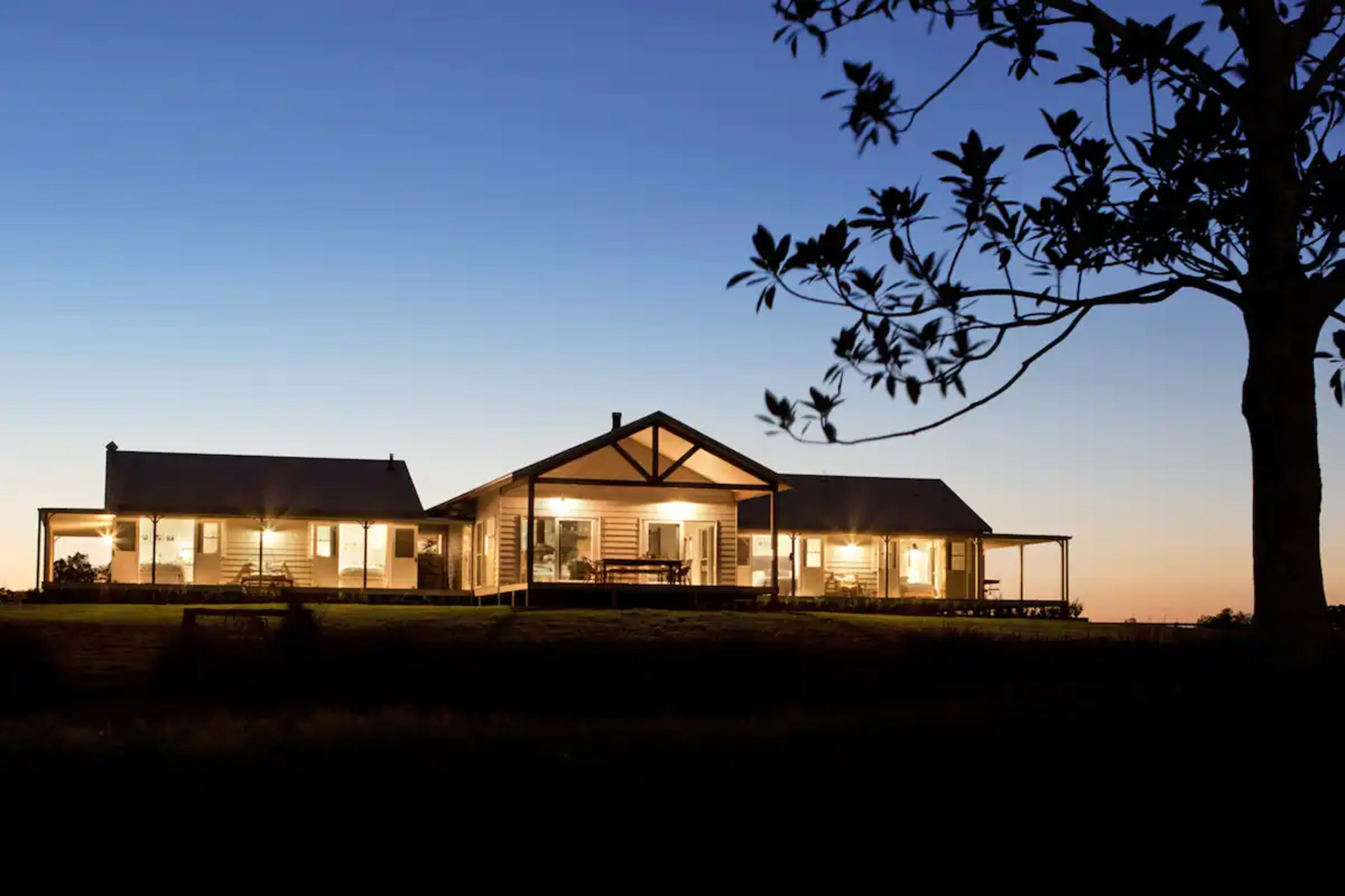 A large house with a veranda is illuminated at dusk, surrounded by trees and an open field.