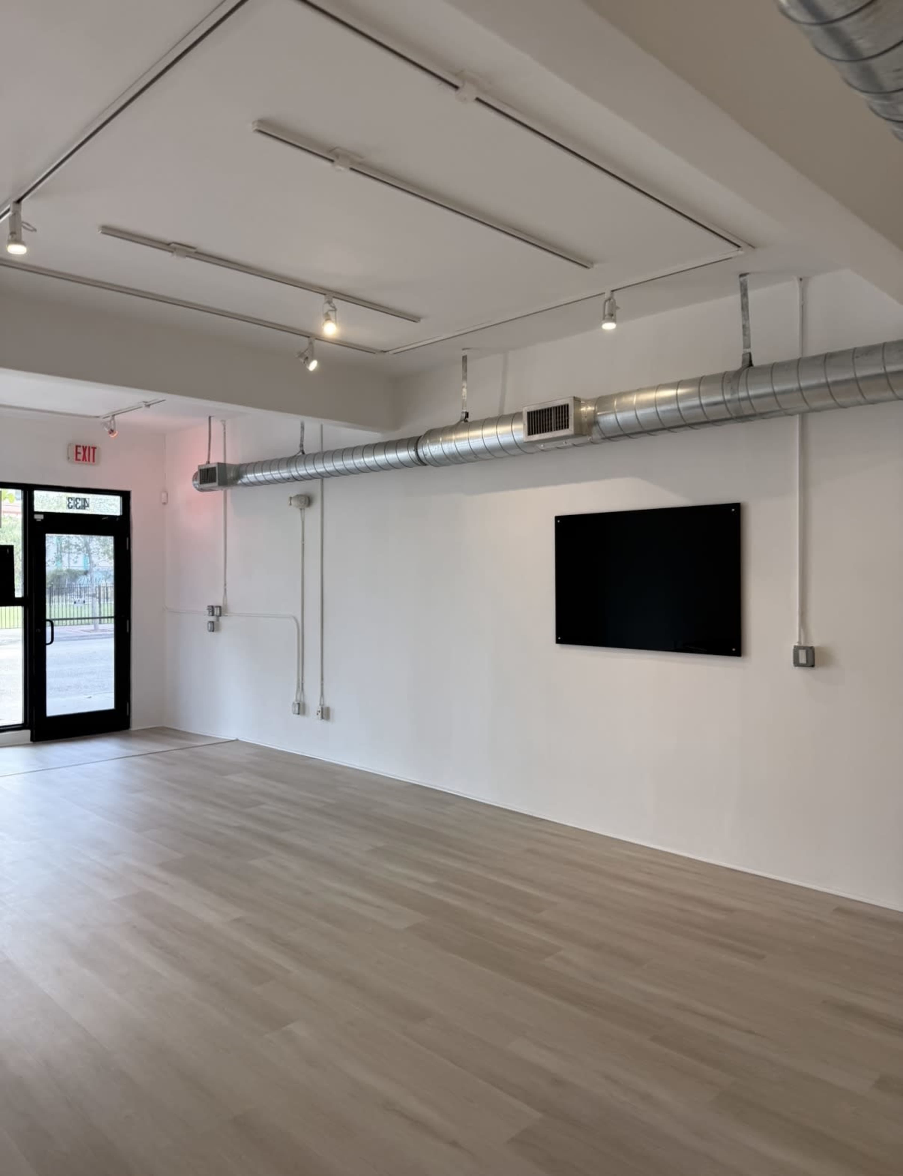The image shows a spacious, empty room with white walls, wooden flooring, a wall-mounted television, and a large window featuring a glass door.