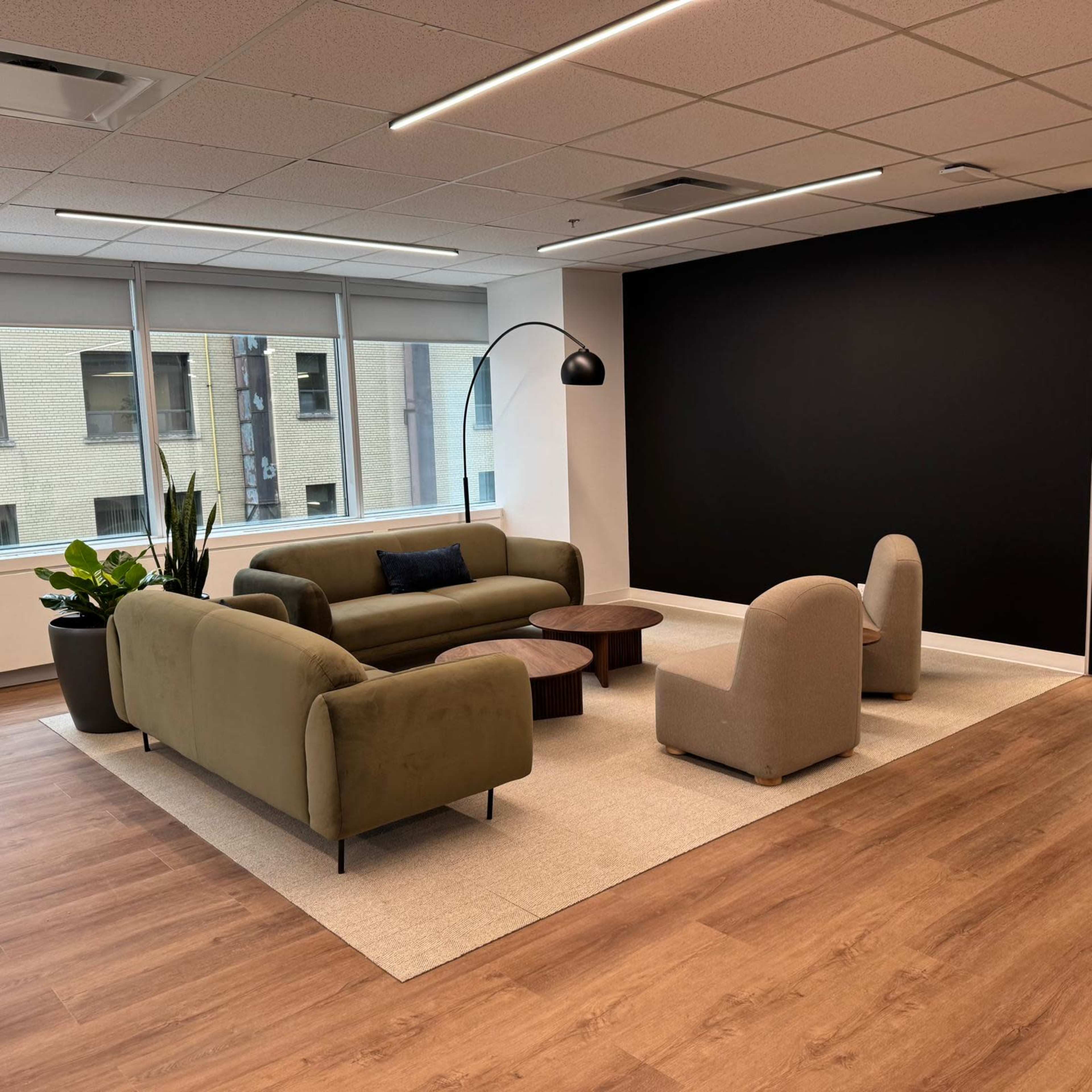 The image shows a modern office lounge area with a green sofa, two light-colored chairs, a wooden coffee table, and a black wall.