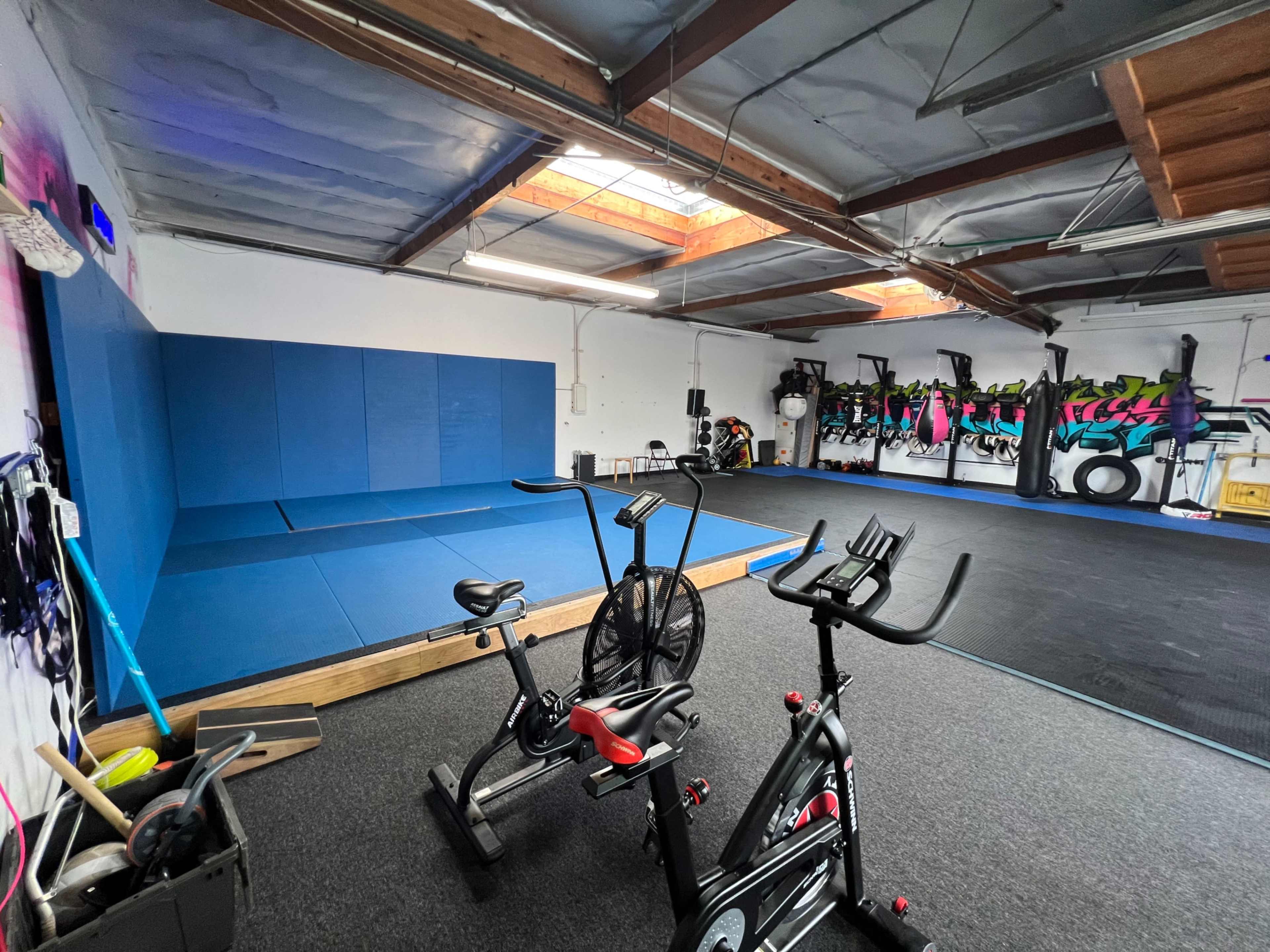 An indoor gym with blue mats on one wall, exercise bikes in the foreground, and various fitness equipment on the opposite wall.