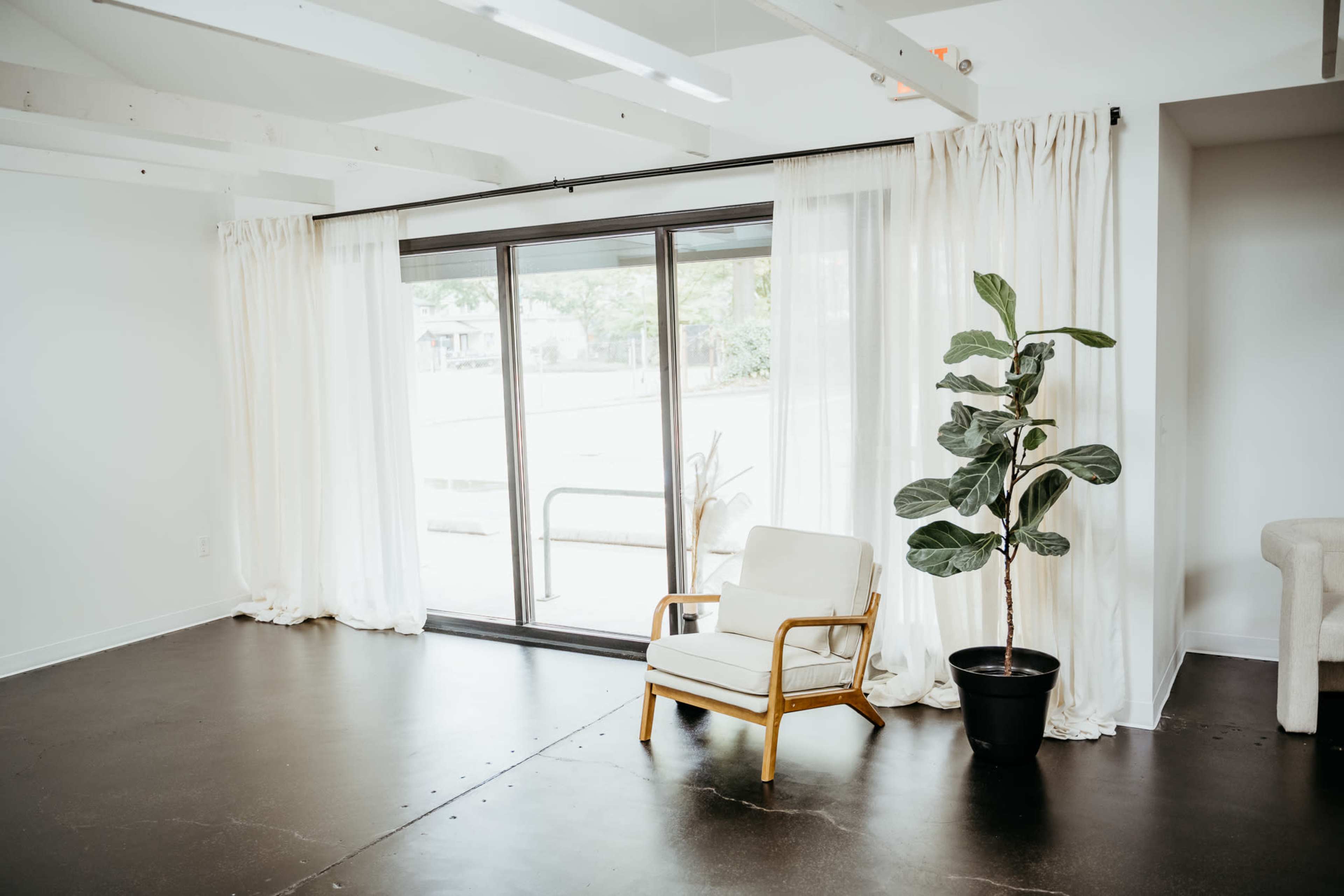 A light-filled room features a white chair with wooden legs beside a large window covered with sheer curtains, along with a potted plant in the corner.