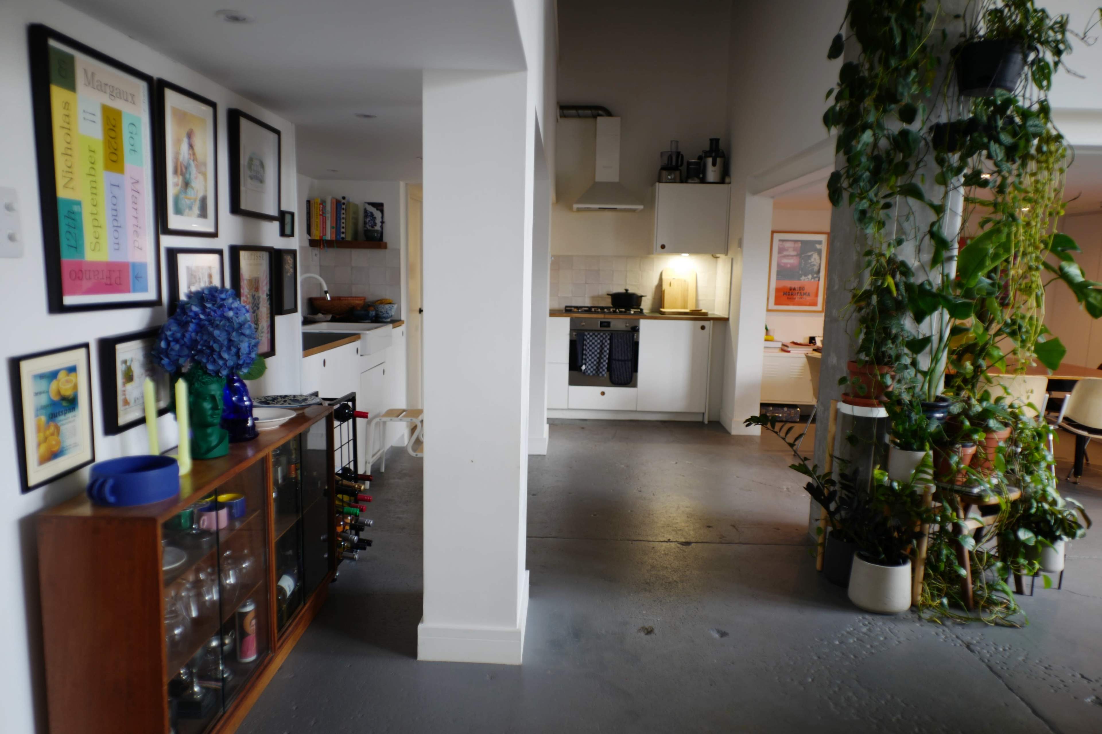 The image shows a spacious kitchen and dining area with a decorative plant-covered column, framed artwork on the walls, and a wooden cabinet filled with glassware.