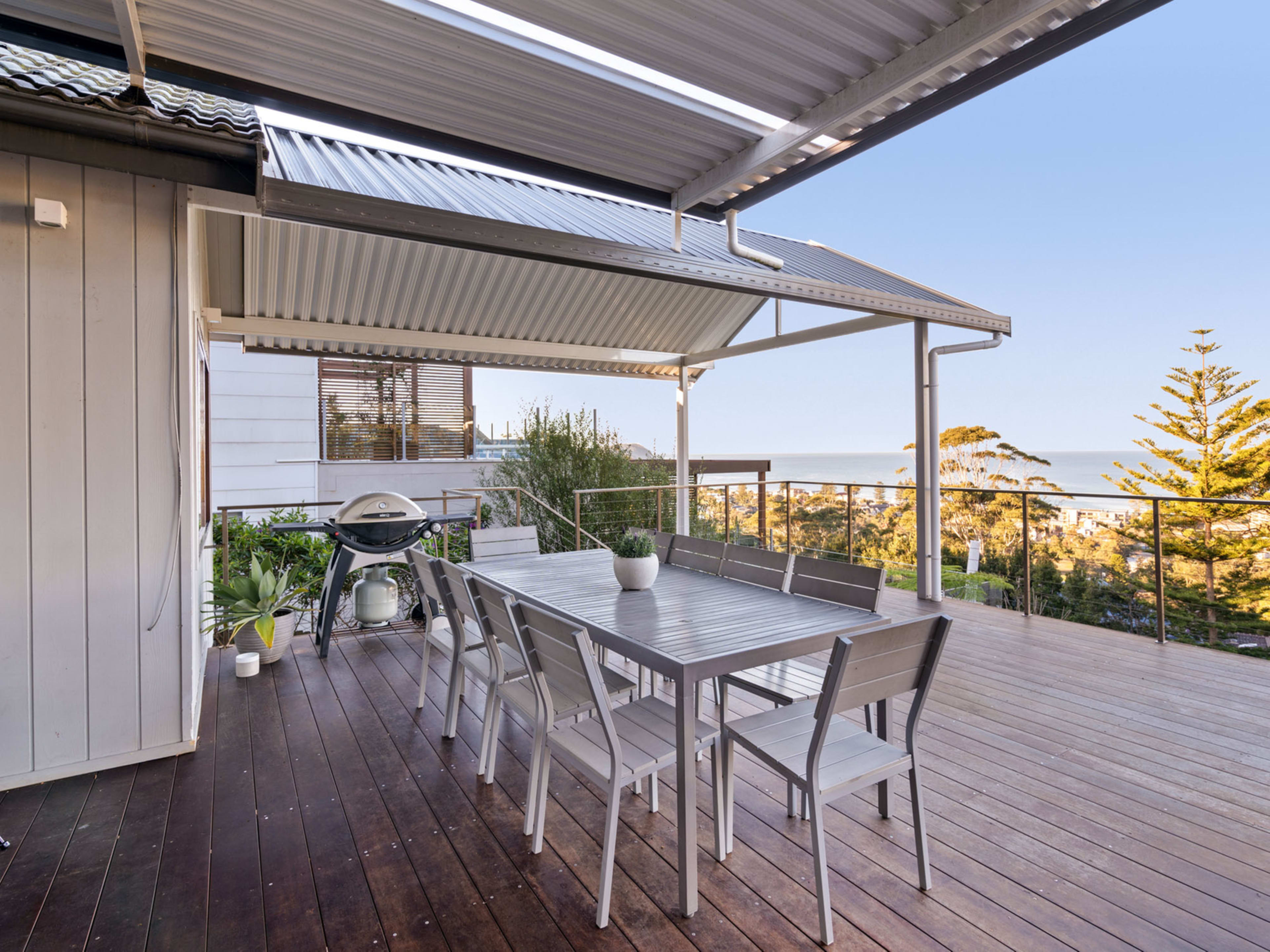 A spacious outdoor deck features a large wooden dining table surrounded by chairs, with a view of the ocean in the distance.