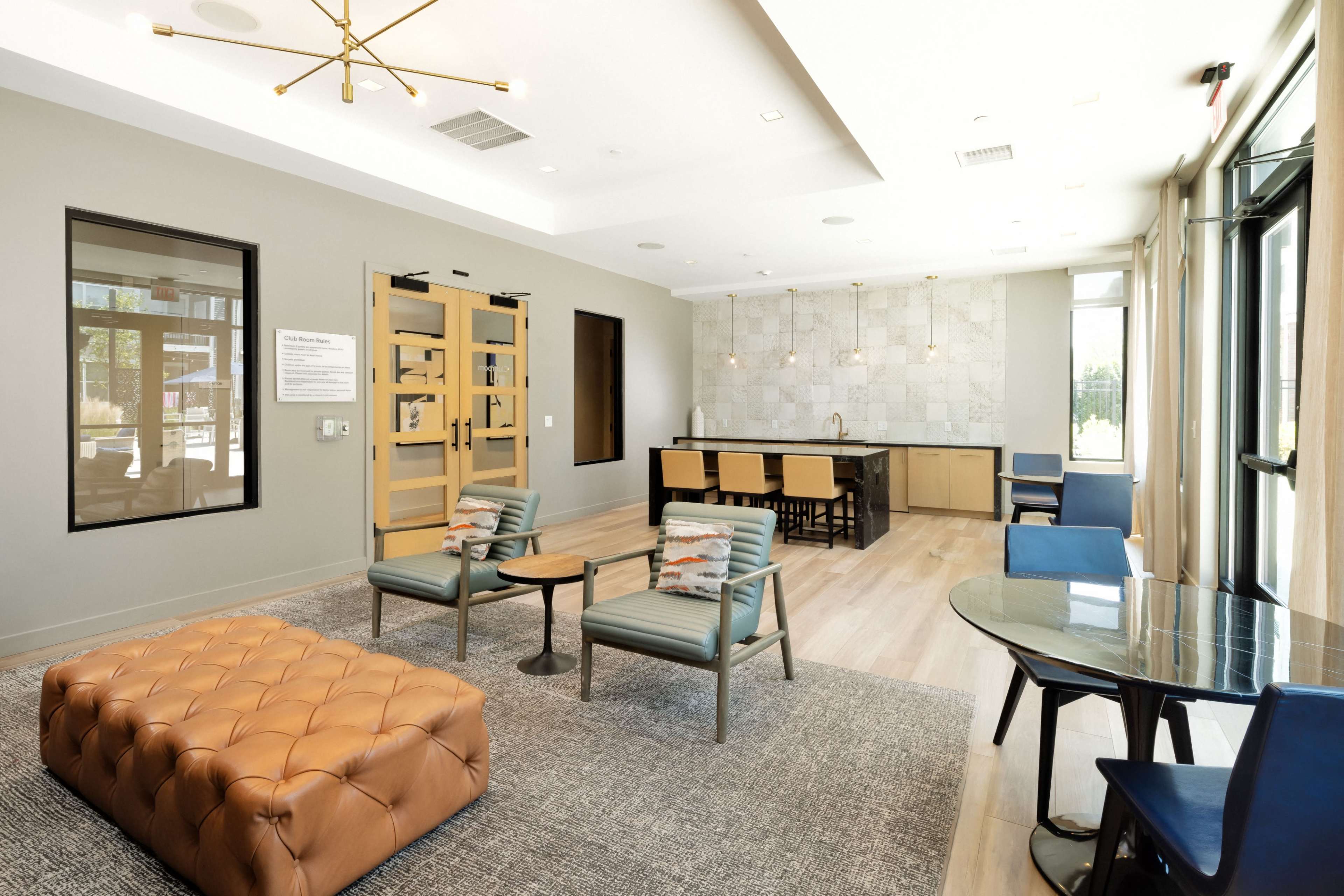 A modern lounge area with a tufted brown ottoman, seating arrangements, and a kitchen bar in a brightly lit space featuring large windows.