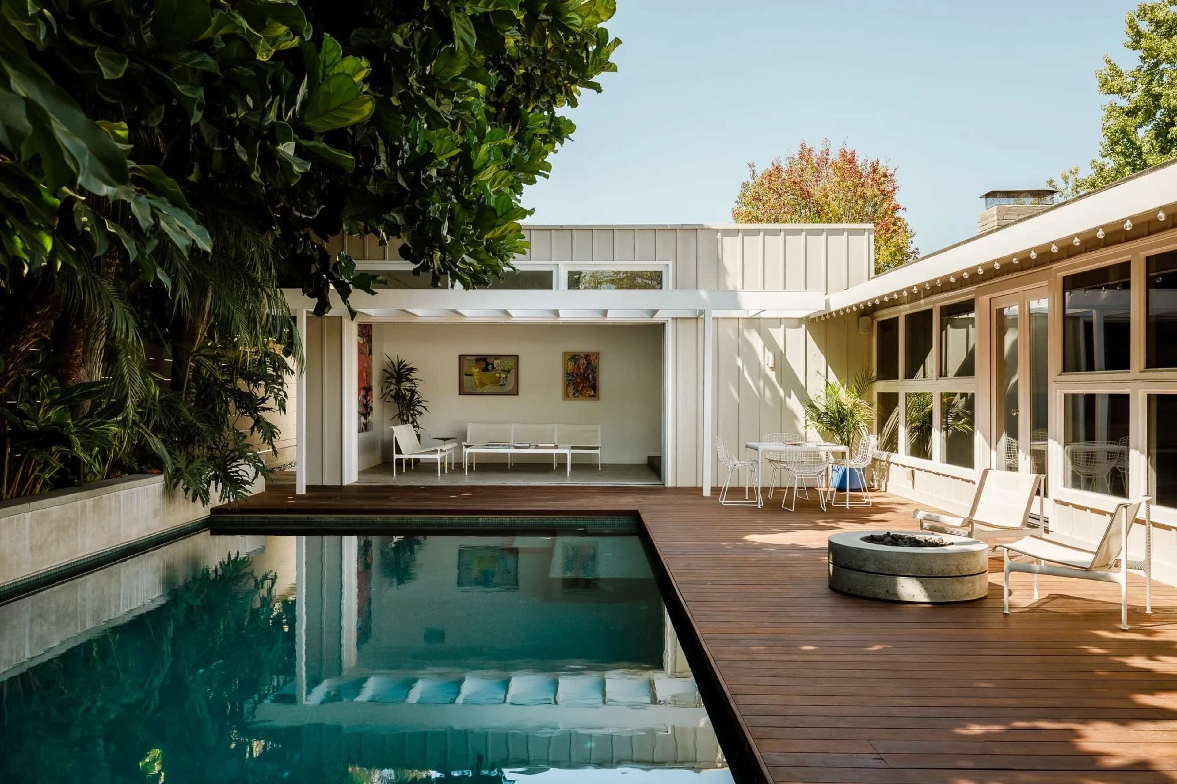 A modern patio features a swimming pool surrounded by wooden decking and lush greenery, with seating areas and large windows in the background.