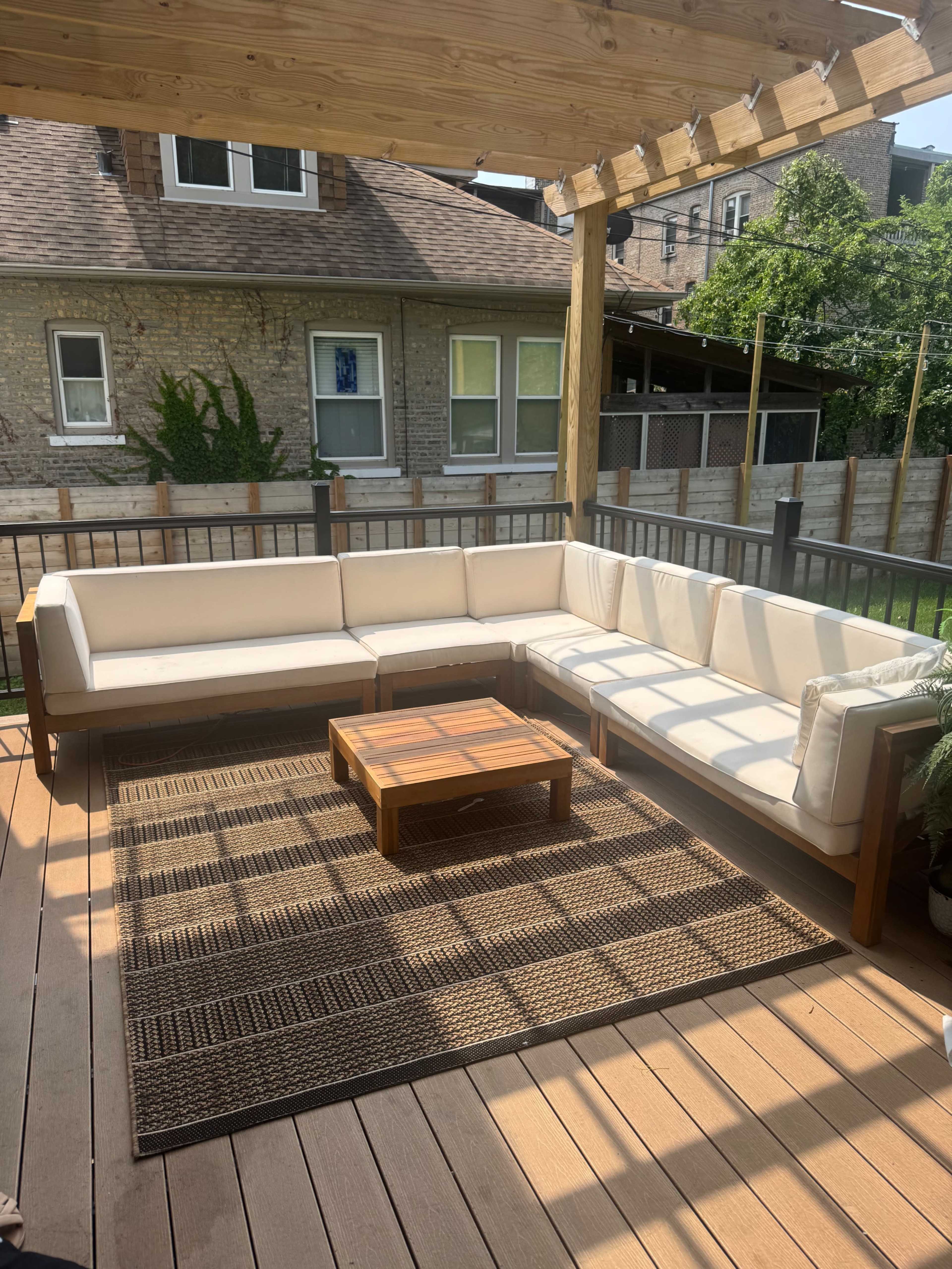 A wooden patio features a sectional sofa with white cushions and a wooden coffee table on a woven rug.