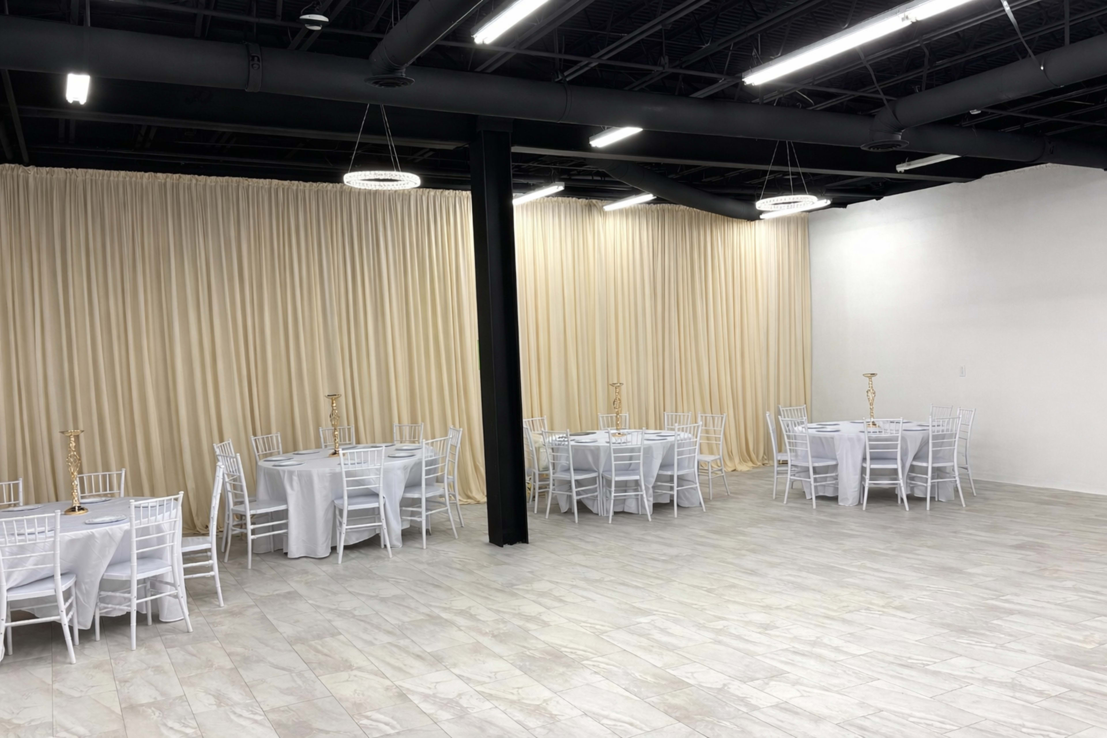 A spacious event room features multiple round tables with white tablecloths and white chairs, separated by a backdrop of beige curtains.