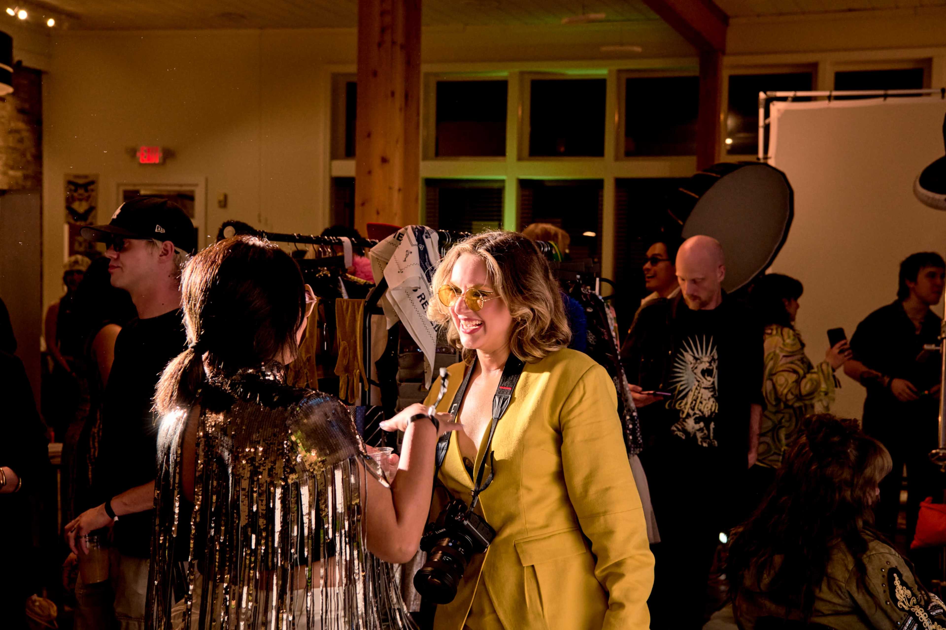 A woman in a yellow outfit smiles while talking to another person wearing a fringed jacket in a crowded, well-lit indoor event.