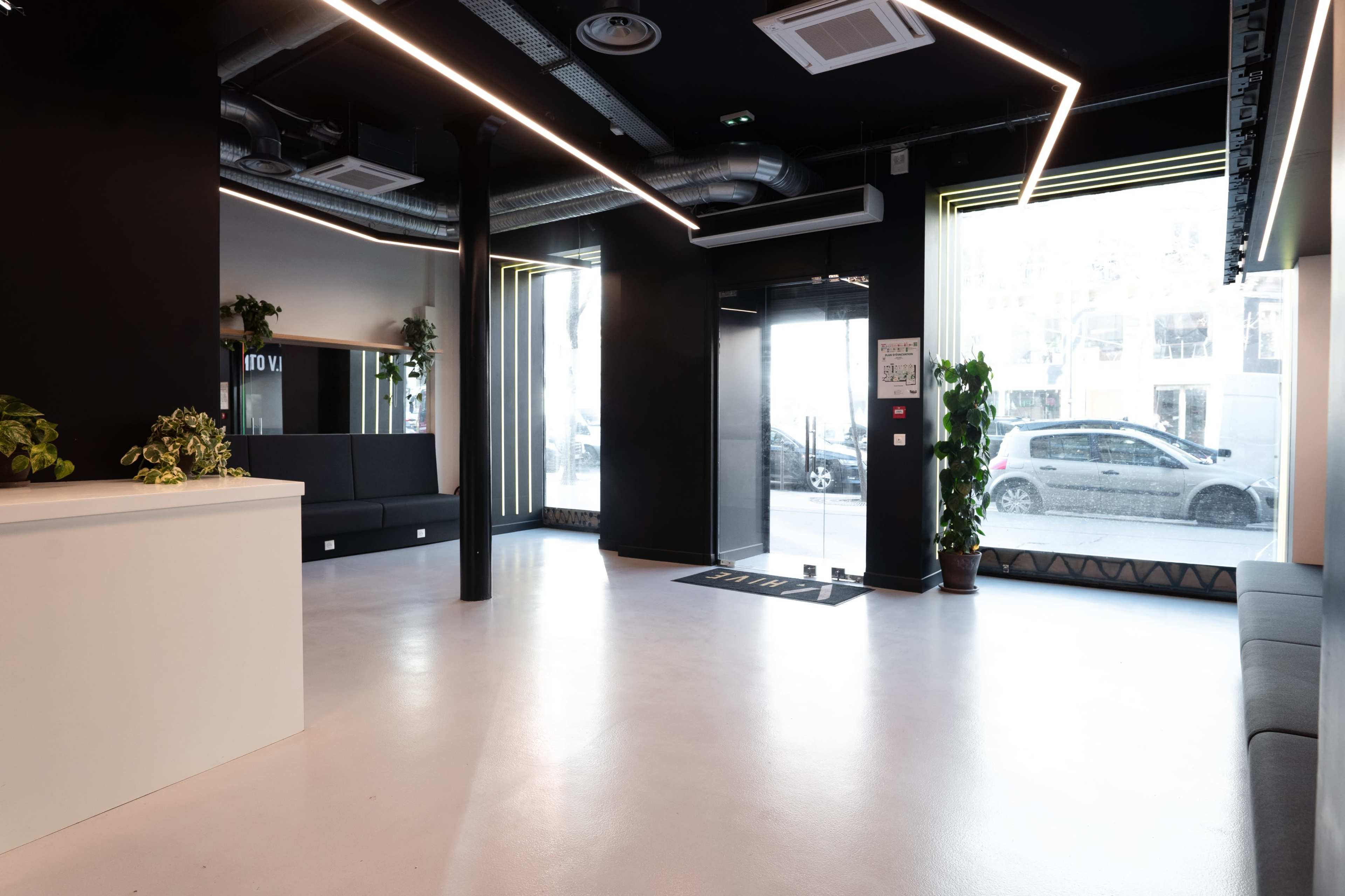 The image depicts a spacious, modern lobby with large windows, a reception area, and plants positioned around the room.